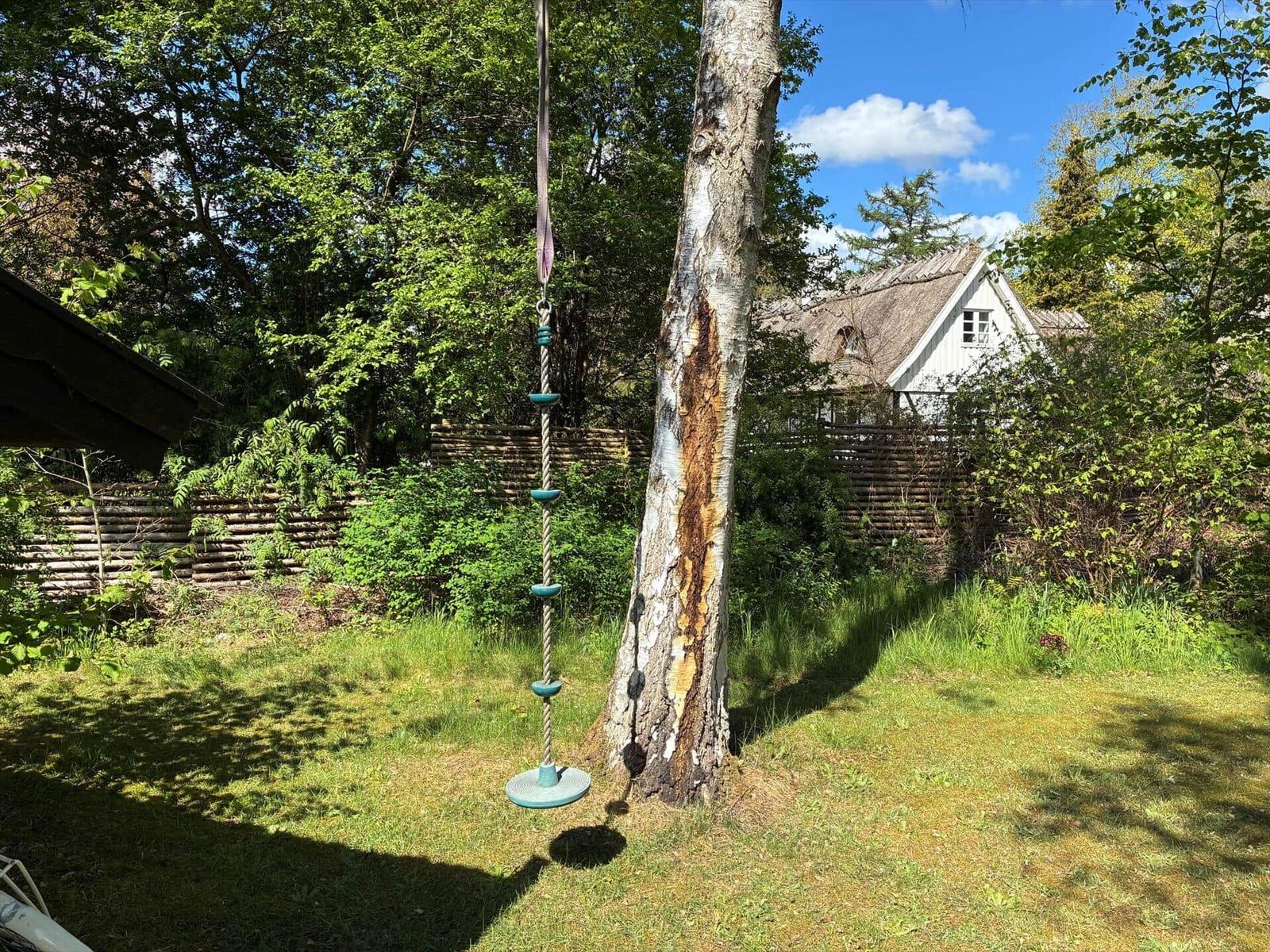 Ein Klettergerüst hängt an einem Baum im Garten. Im Hintergrund ist ein Haus mit Schindeldach sichtbar.