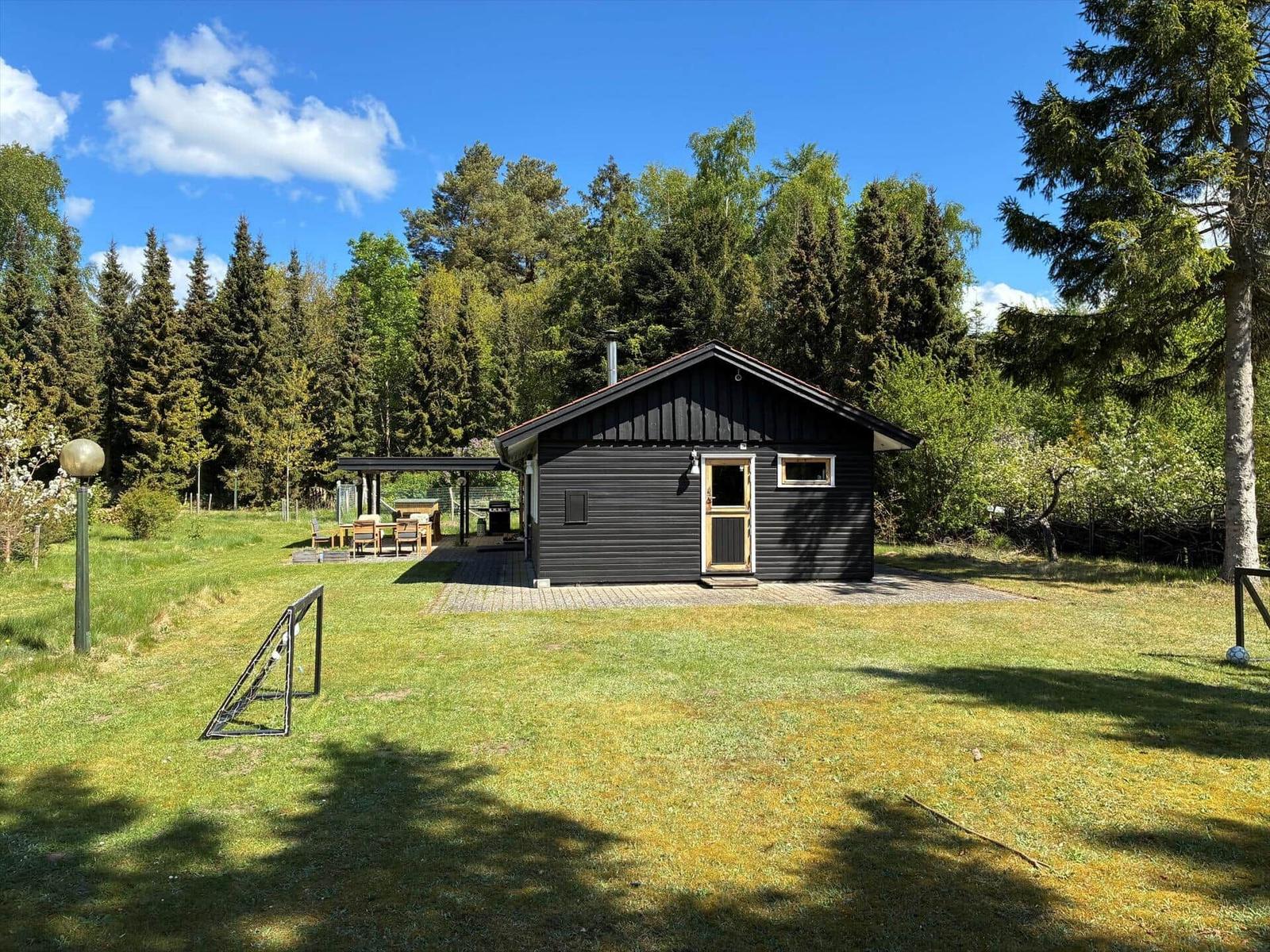 Schwarze Hütte mit Terrasse und Grill, umgeben von Gras und Bäumen unter blauem Himmel.