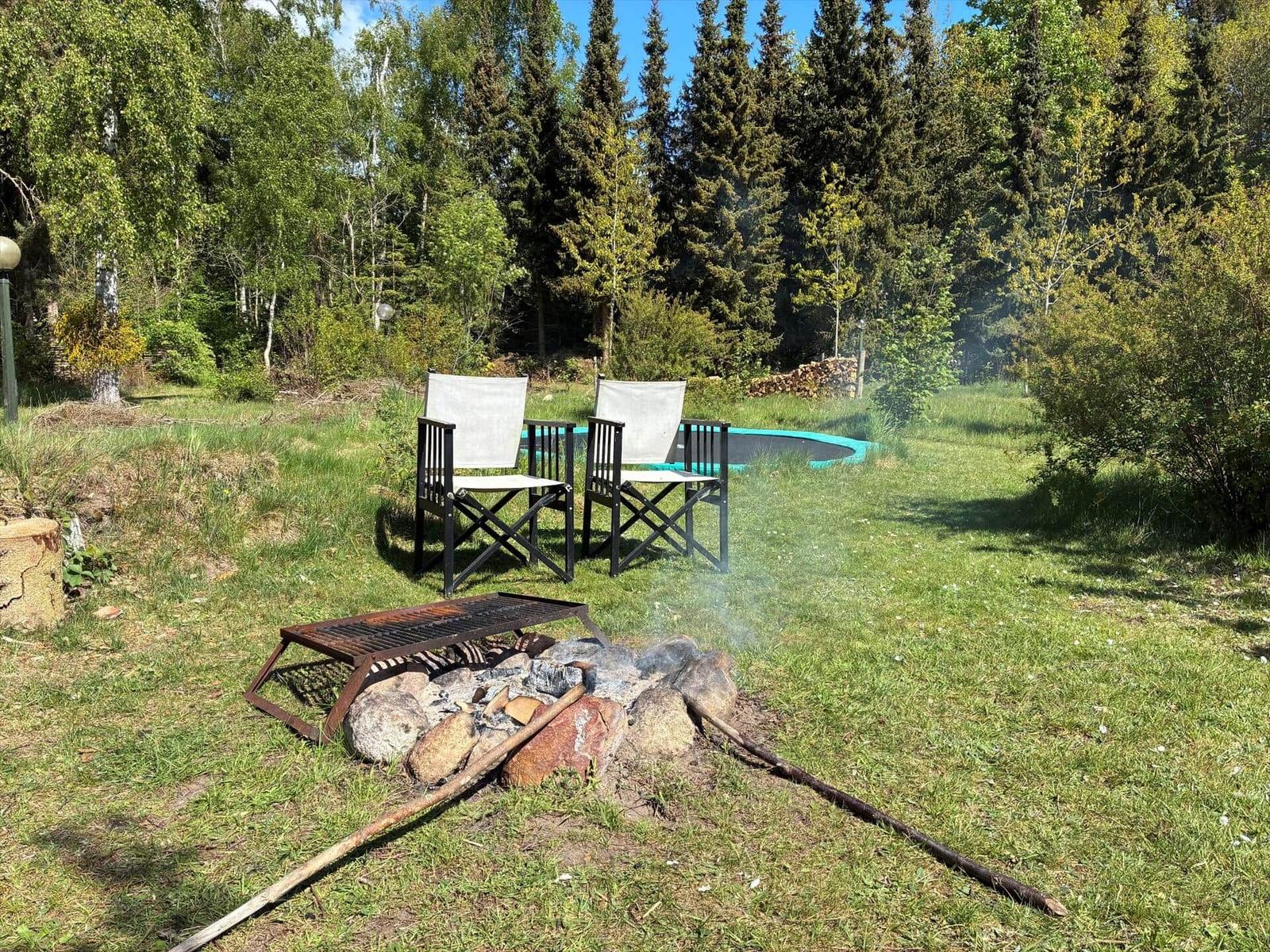 Feuerstelle mit Grill und zwei Stühlen im Gras, Hintergrund mit Wald und Sprungbad.