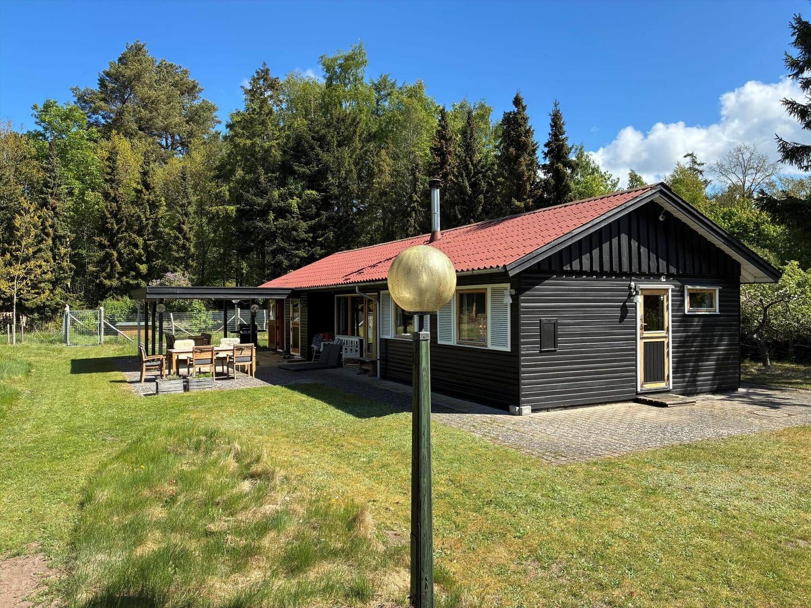 Schwarze Holzhütte mit rotem Dach, Garten und Terrasse unter Bäumen.