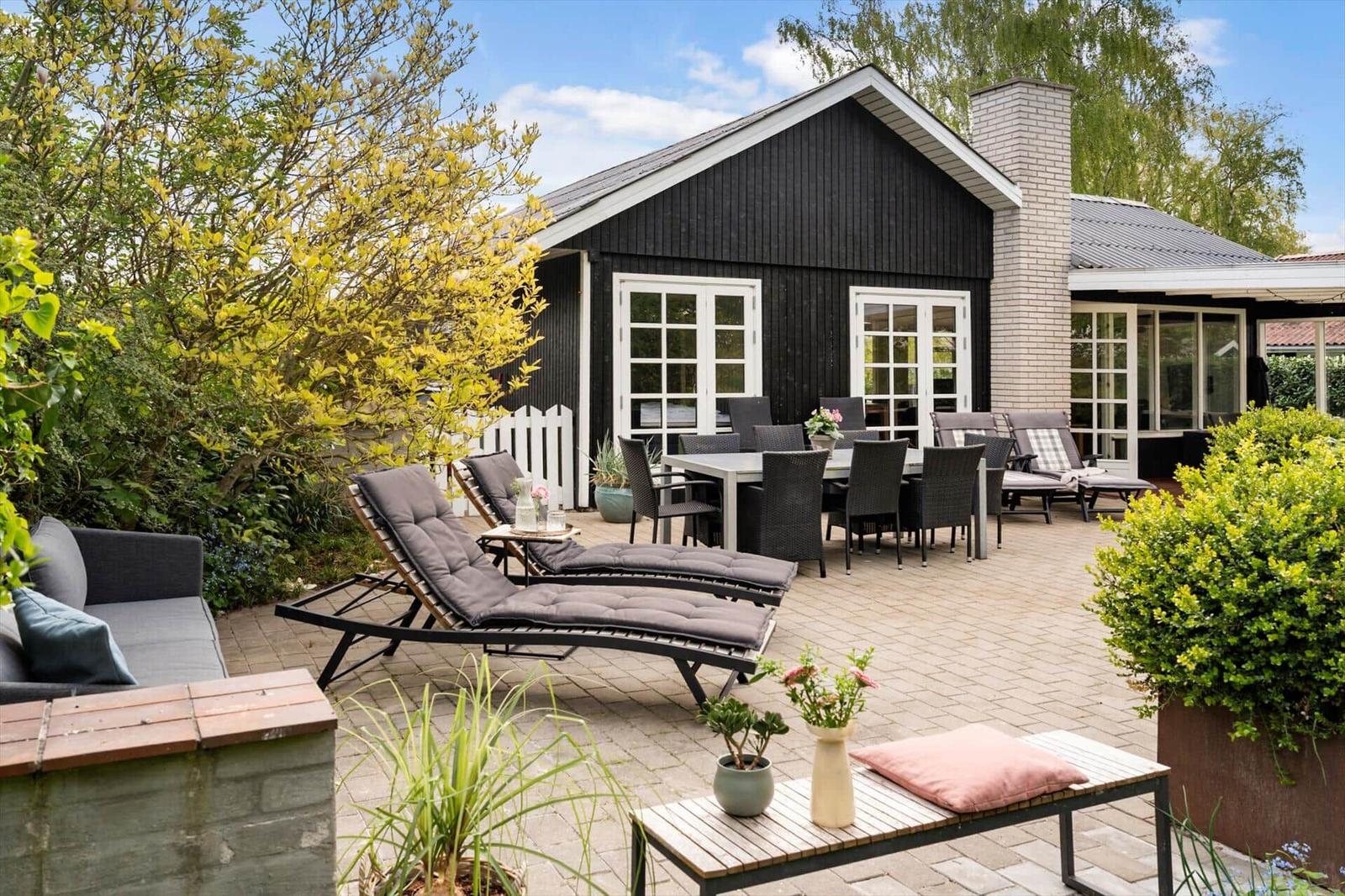 Hinterhof mit Liegestühlen, Tisch und Gartenmöbeln vor schwarzem Haus mit weißen Fenstern.