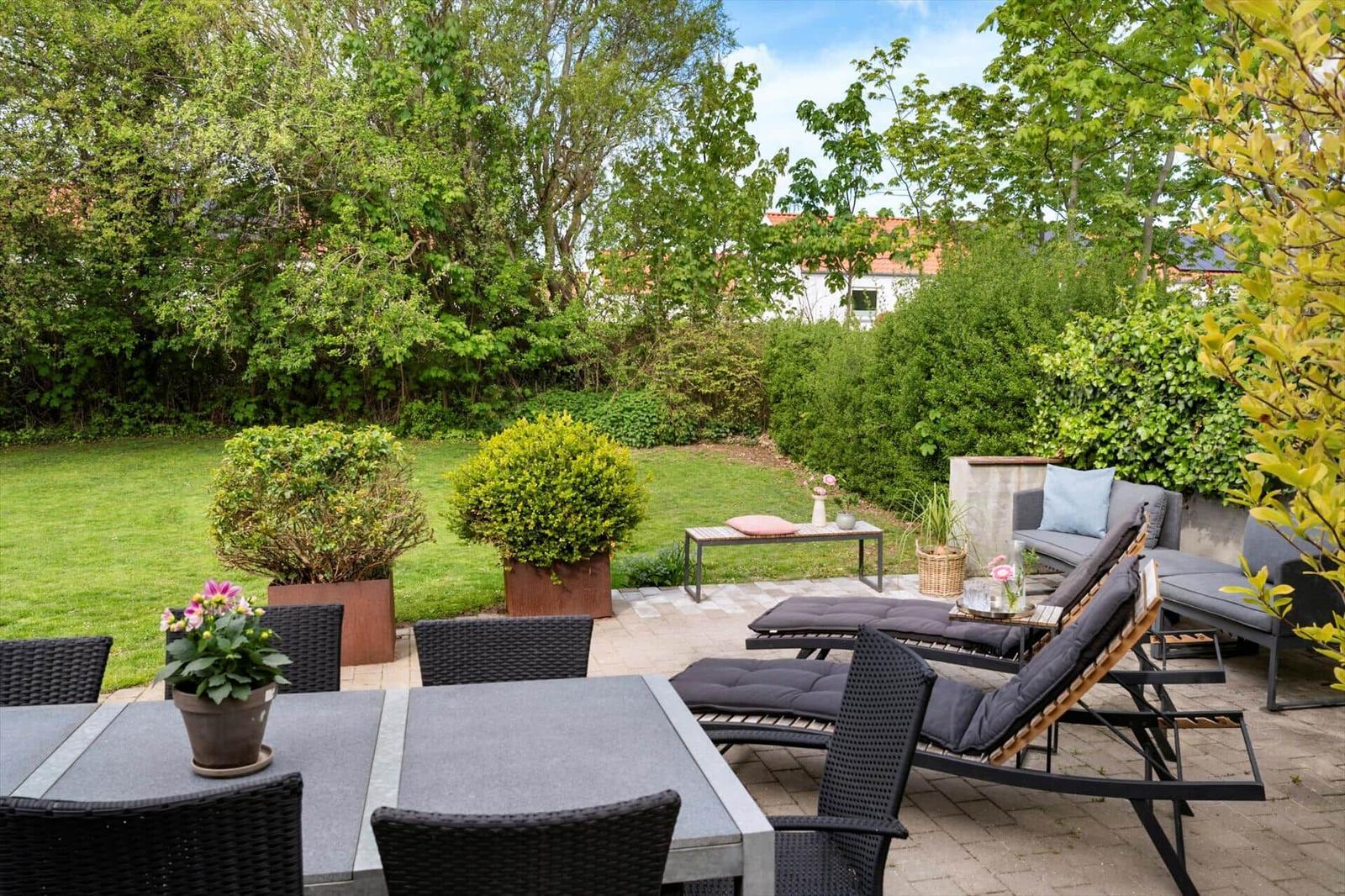 Terrasse mit Gartenmöbeln, Tisch, Stühlen und Pflanzen. Grünfläche und Bäume im Hintergrund.