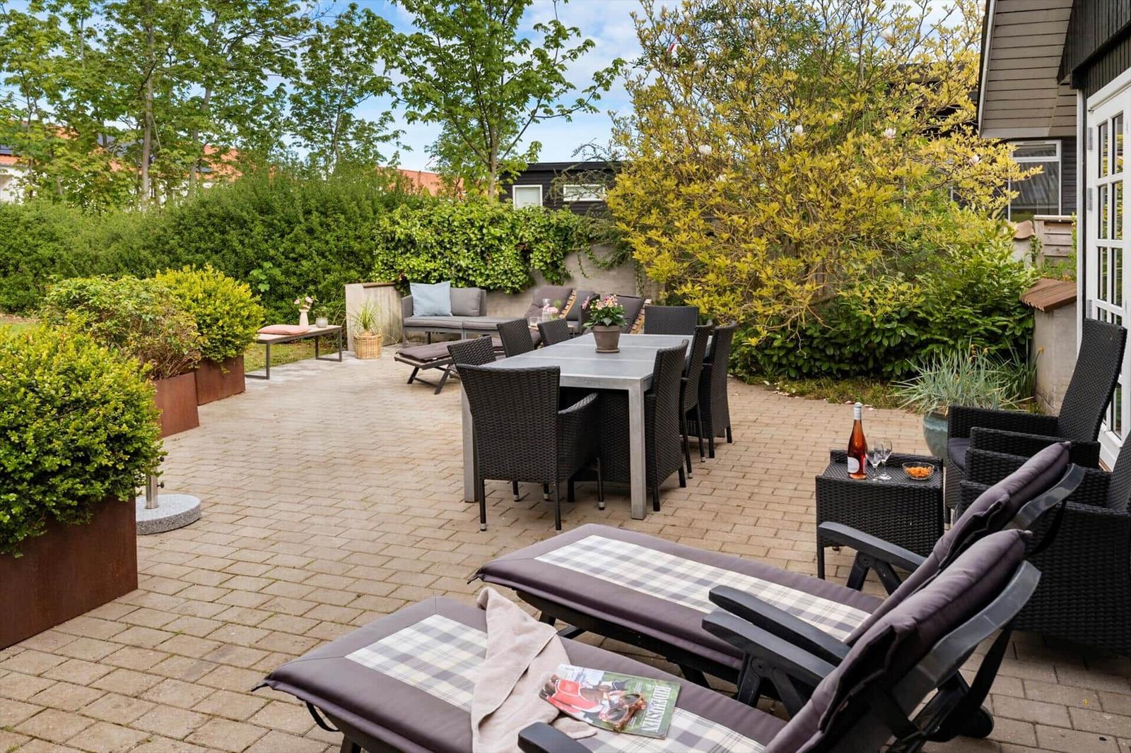 Grosser gepflasterter Garten mit Sitzgruppe, Tisch und Liegestühlen. Umgeben von Bäumen und Sträuchern.