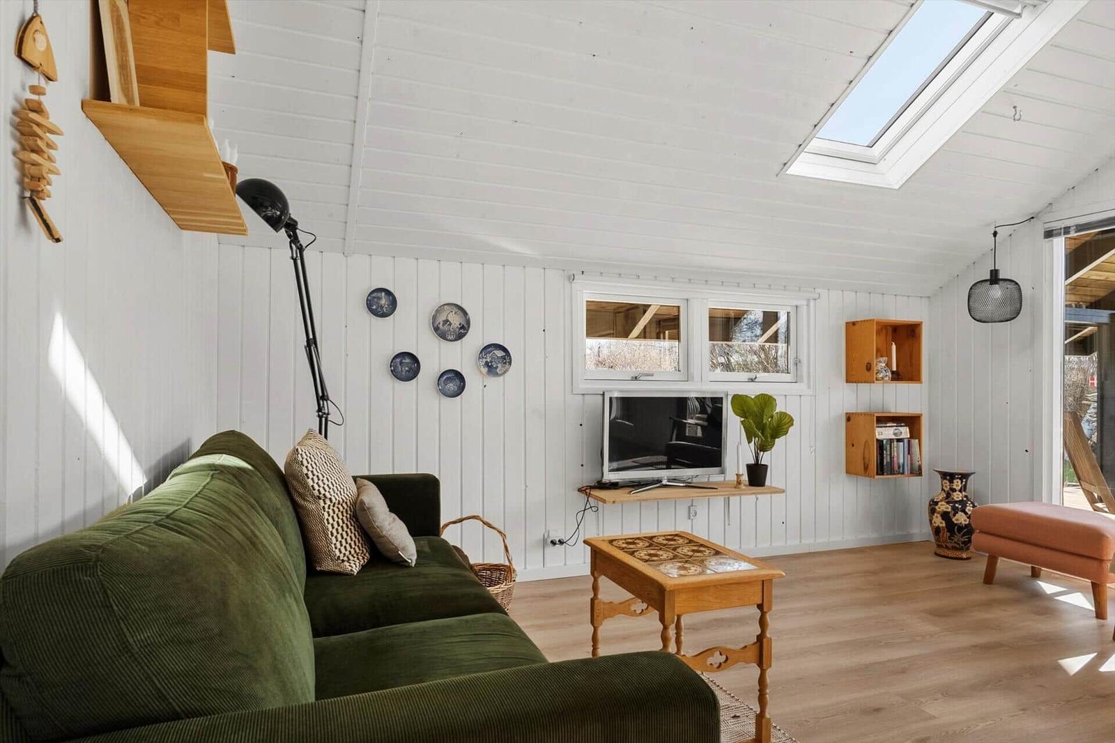 Wohnzimmer mit grünem Sofa, Holztisch und TV unter Fenster. Decke mit Dachfenster und Holzwand.