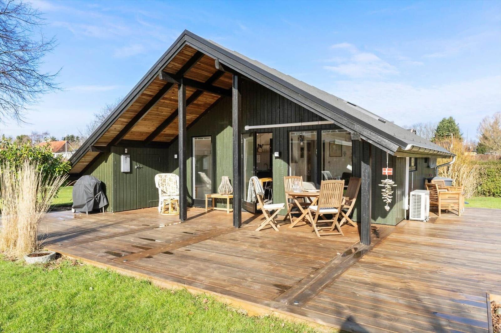 Holzterrasse mit Sitzgelegenheiten vor einem grünen Holzhaus mit großen Fenstern.