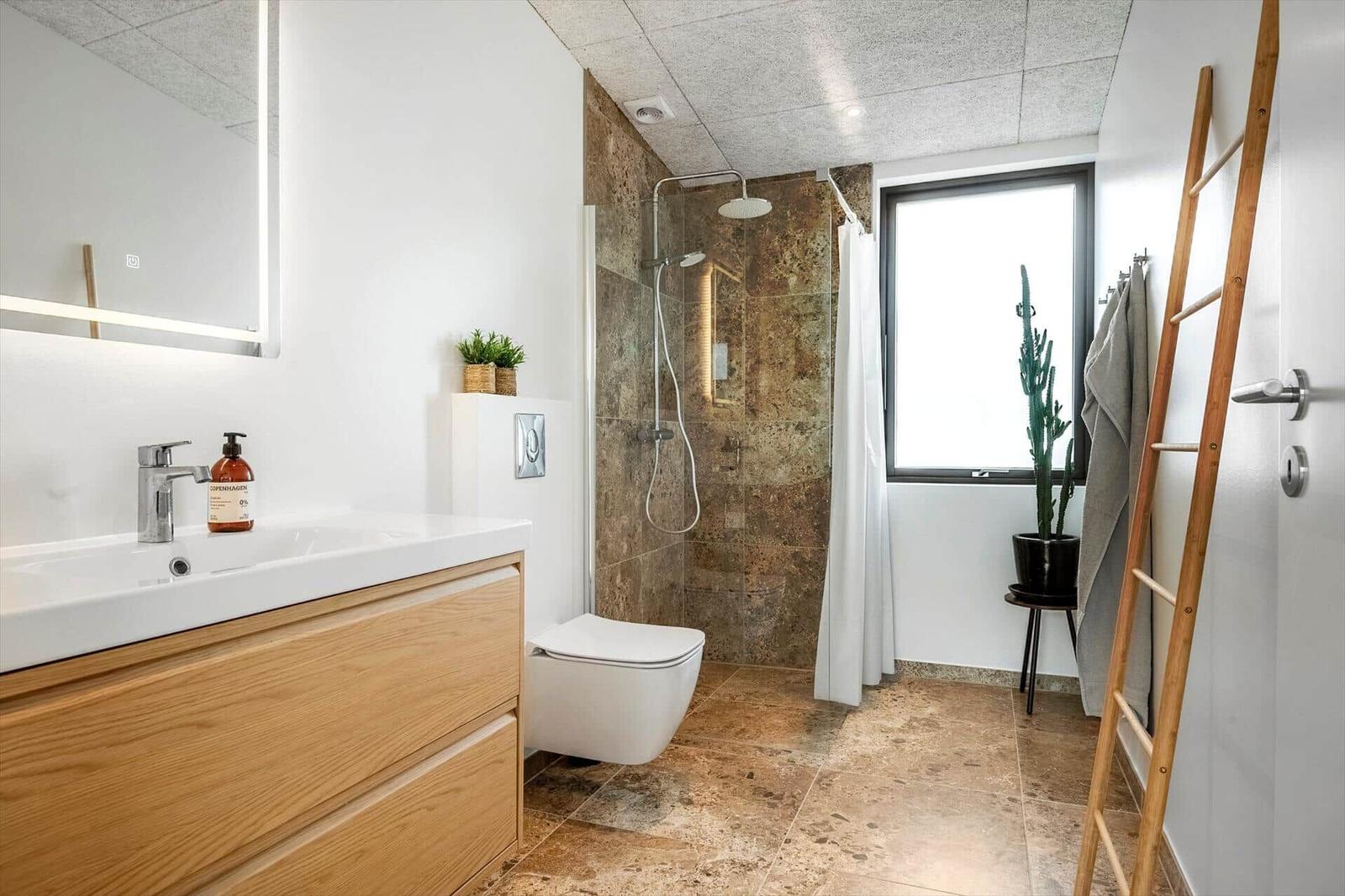 Badezimmer mit Dusche, Waschbecken und Toilette. Holzwaschtisch, Fenster und Pflanzen.