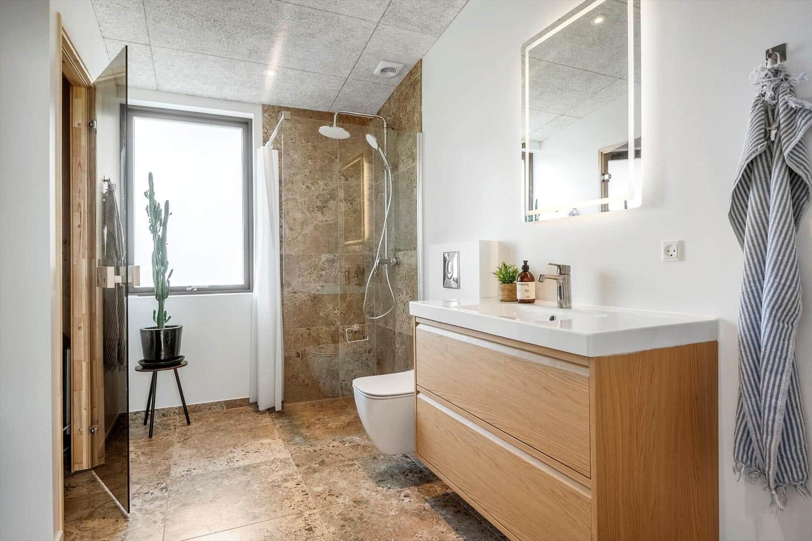 Badezimmer mit Dusche, Waschbecken und Toilette. Holzschrank, Spiegel und Pflanze.