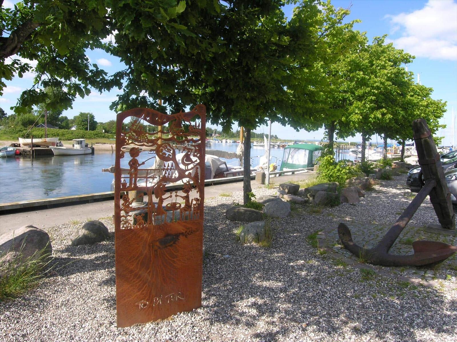 A rustic metal screen and a large anchor lie on gravel by the water.