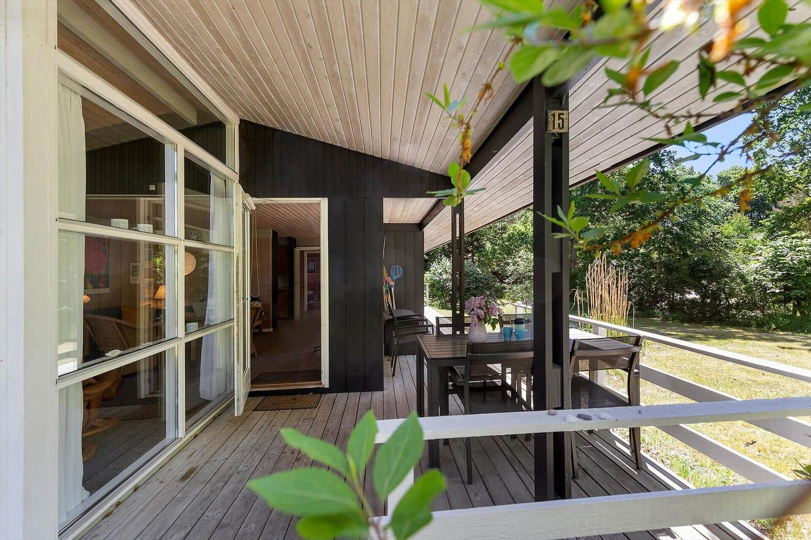 Deck with table and chairs, view into living area, wooden cladding and green surroundings.