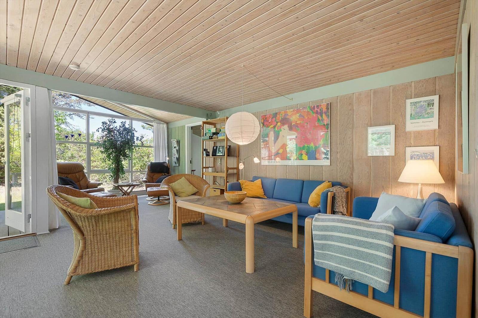 Living room with blue sofas, wooden table and wicker chairs. Wooden wall and ceiling. Glass door to garden.