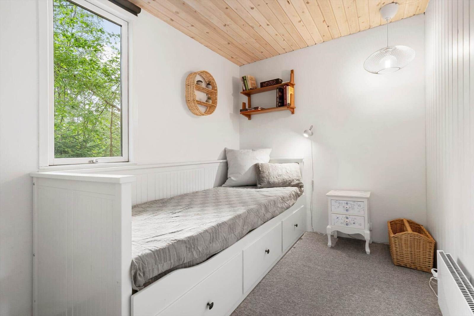 Schlafzimmer mit Bett, Nachttisch und Fenster mit Blick auf Bäume.