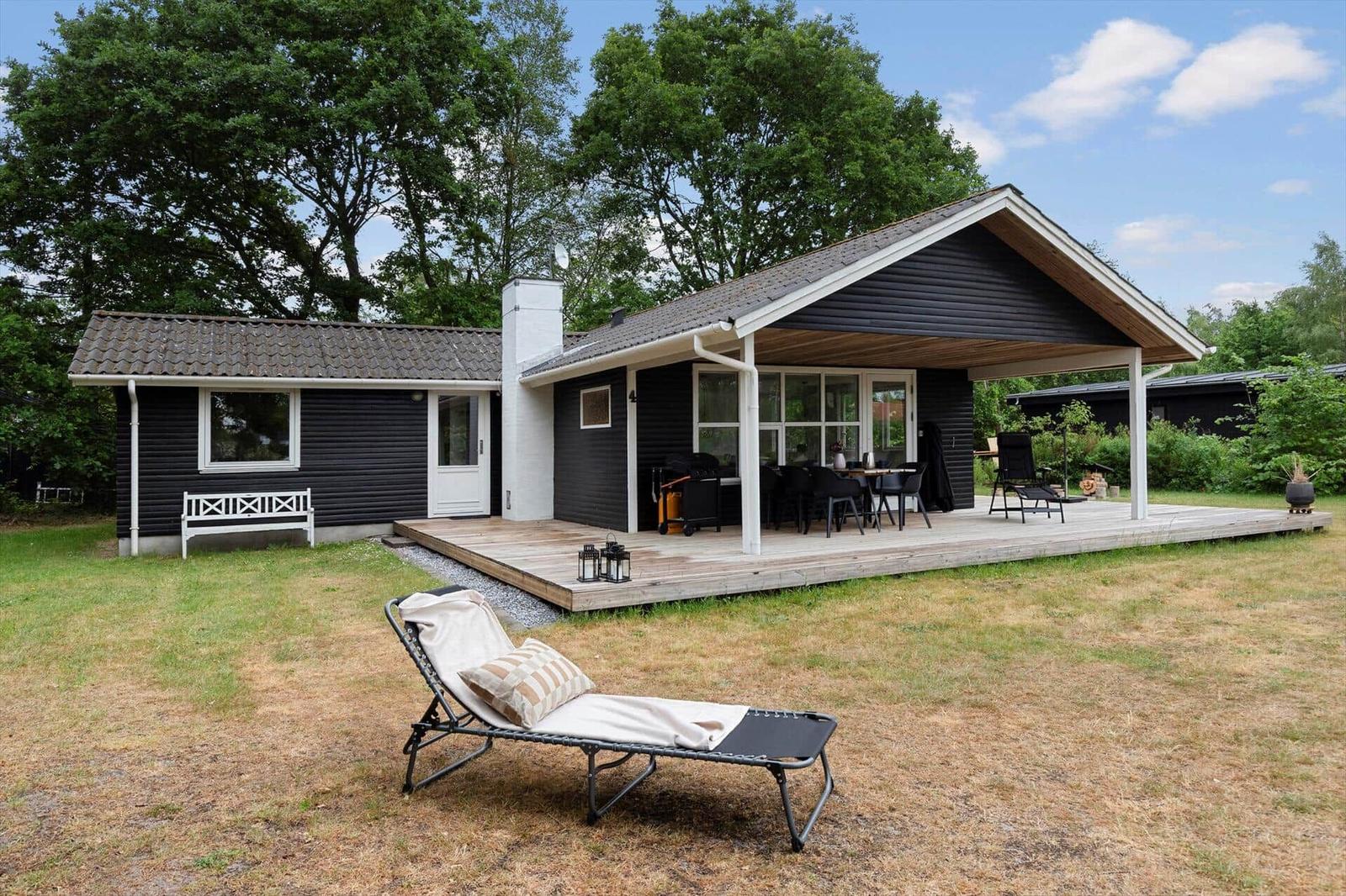 Schwarzes Haus mit Terrasse, Gartenliege und Grillplatz im Grünen.