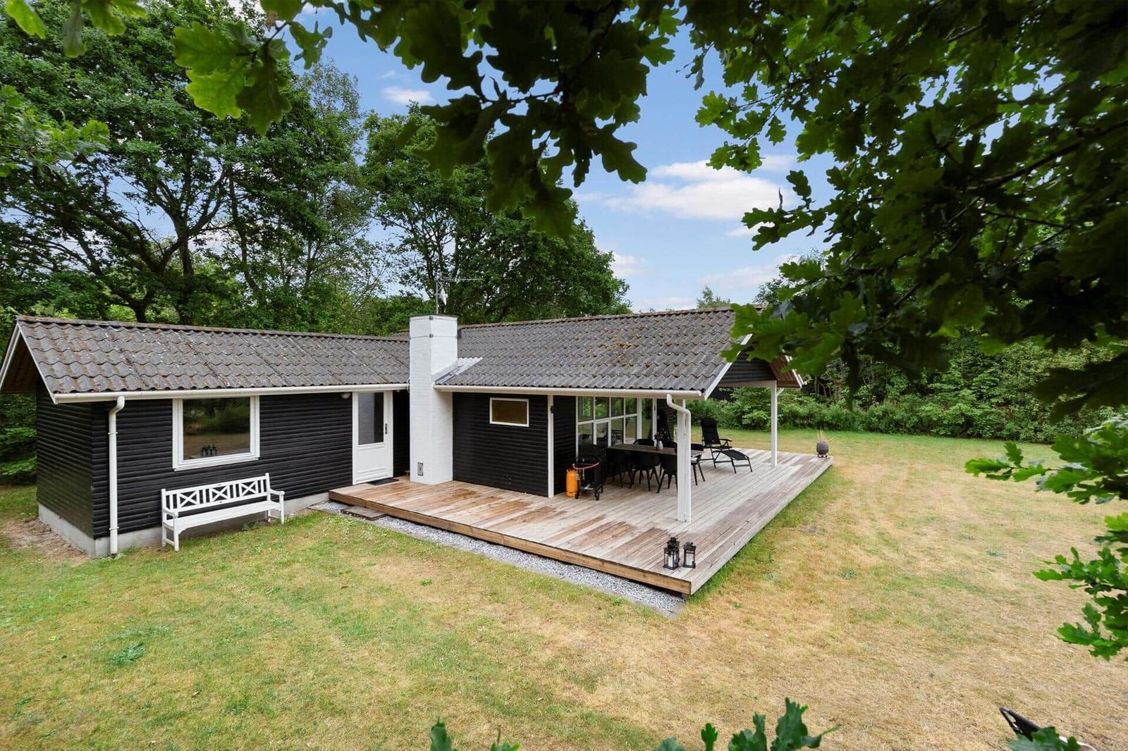 Schwarzes Holzhaus mit Terrasse, Grill und Garten. Blick durch Bäume.