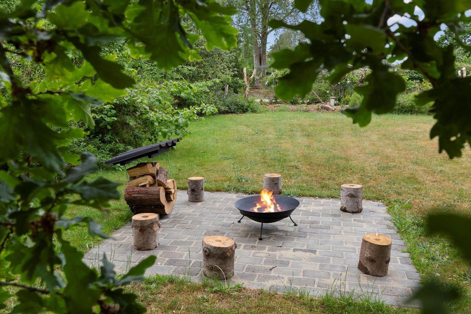 Feuerkorb mit Holzstapel und Sitzbänken im Garten