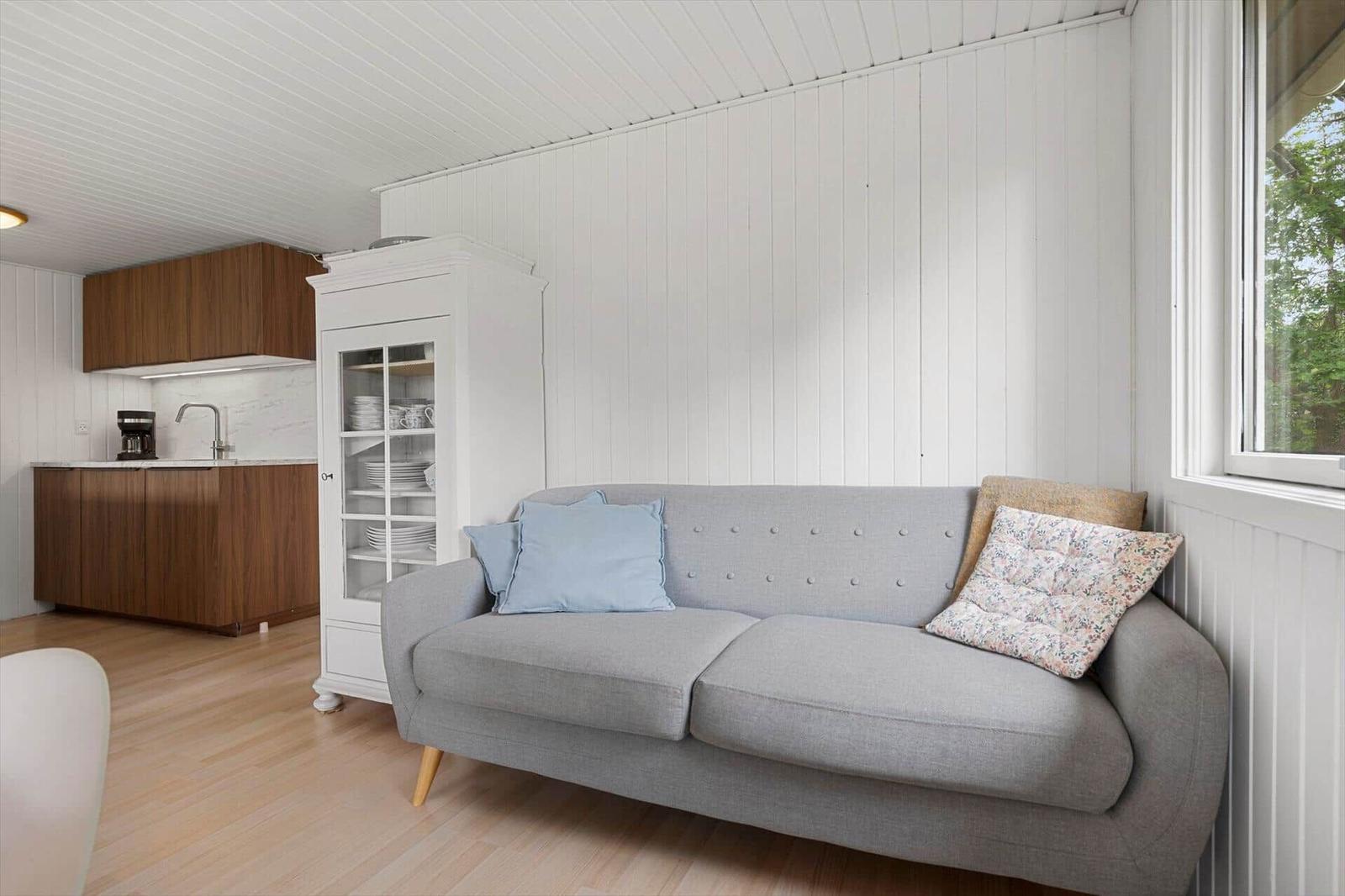 Ein grauer Sofa mit Kissen steht vor einem Fenster. Im Hintergrund ist eine Küche mit Holzmöbeln.