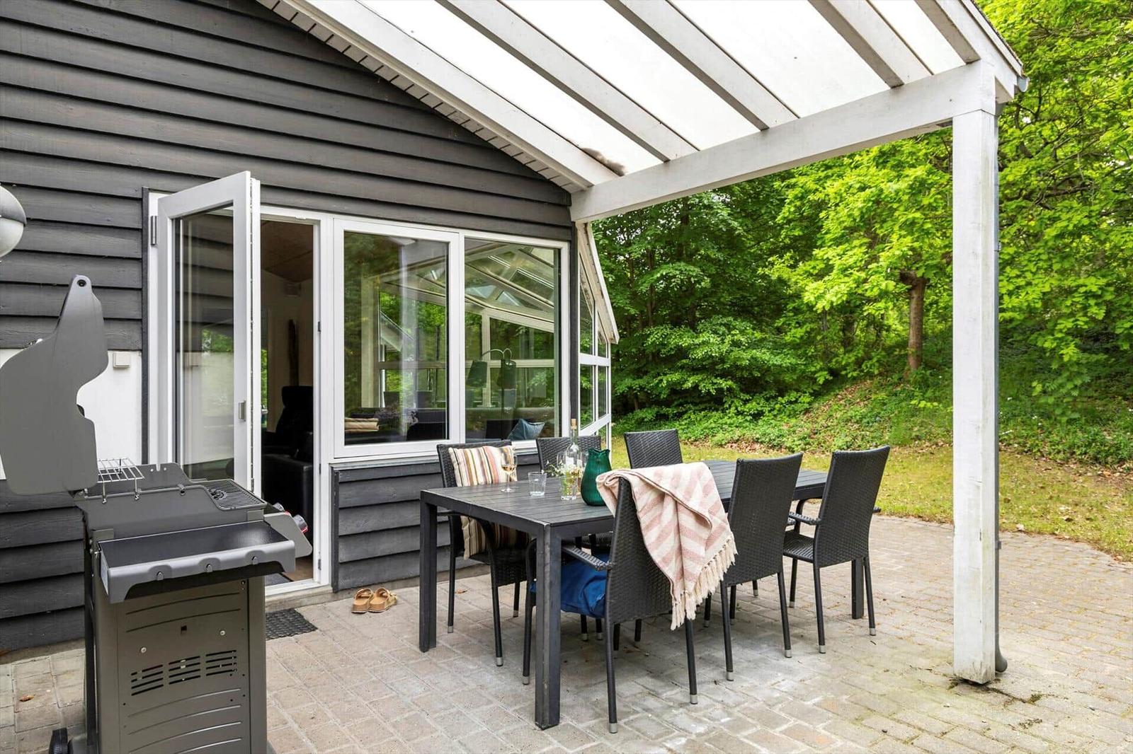 Terrasse mit Tisch, Stühlen und Grill neben Haus. Hintergrund: grüner Wald.