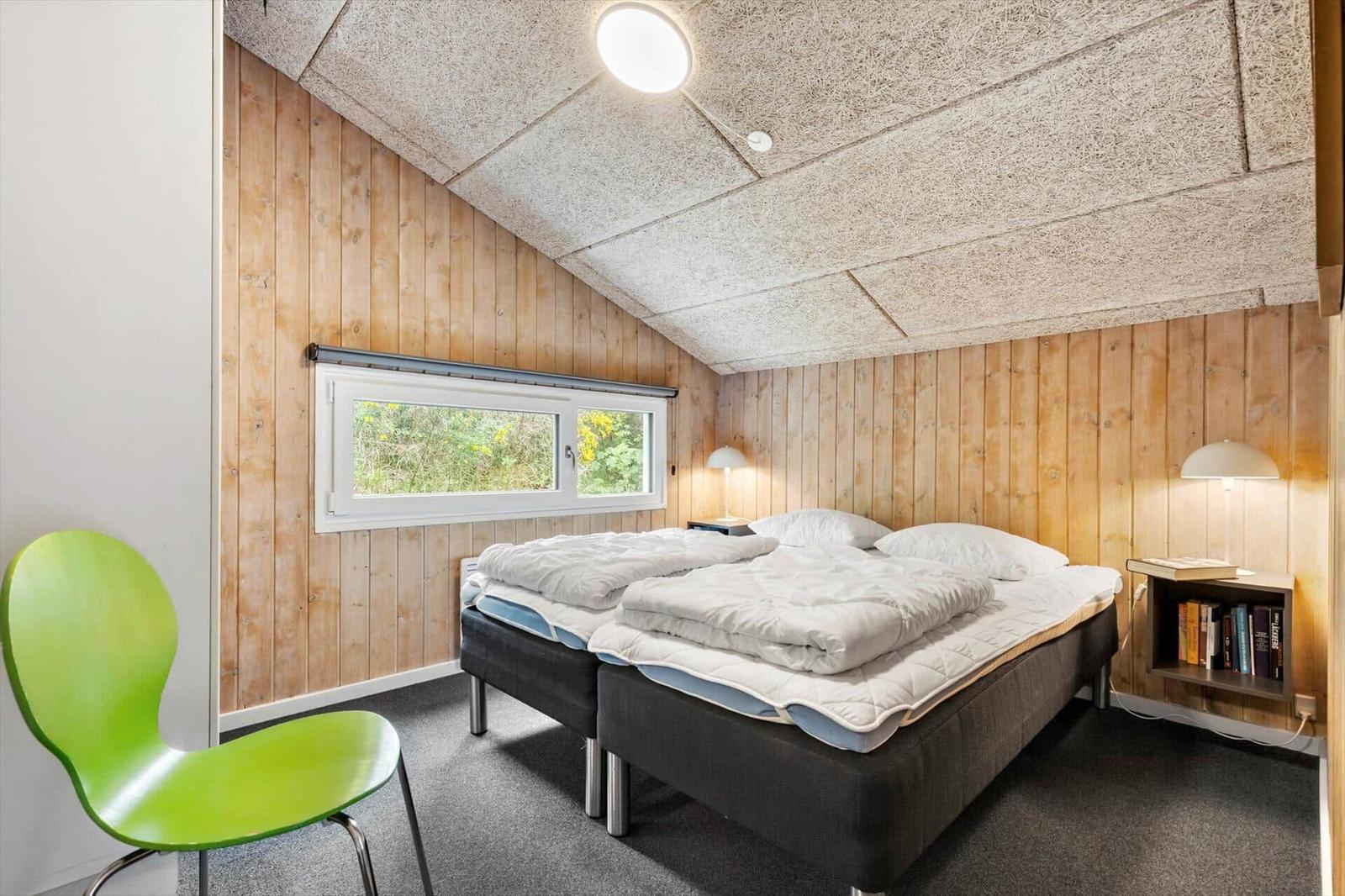 Schlafzimmer mit Doppelbett, grüner Stuhl und Holzwand. Fenster zeigt Außenbereich.