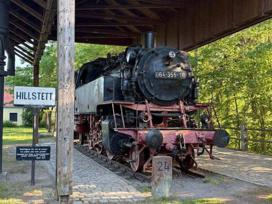 Dampflokomotive 064-355-1 steht auf Bahnsteig unter Dach. Schild zeigt Hillstett. Grün hinter dem Bahnhof.