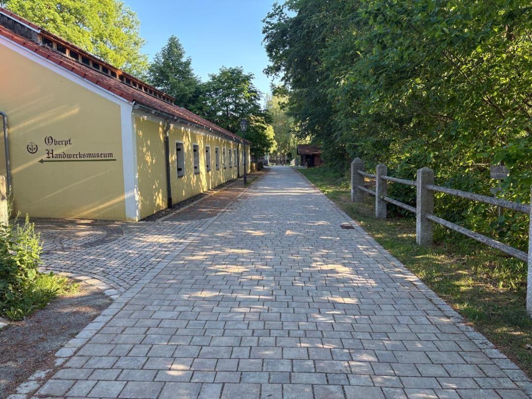 Pflasterweg führt an gelbem Gebäude mit Museumsschild vorbei. Rechts Holzgeländer und Bäume.