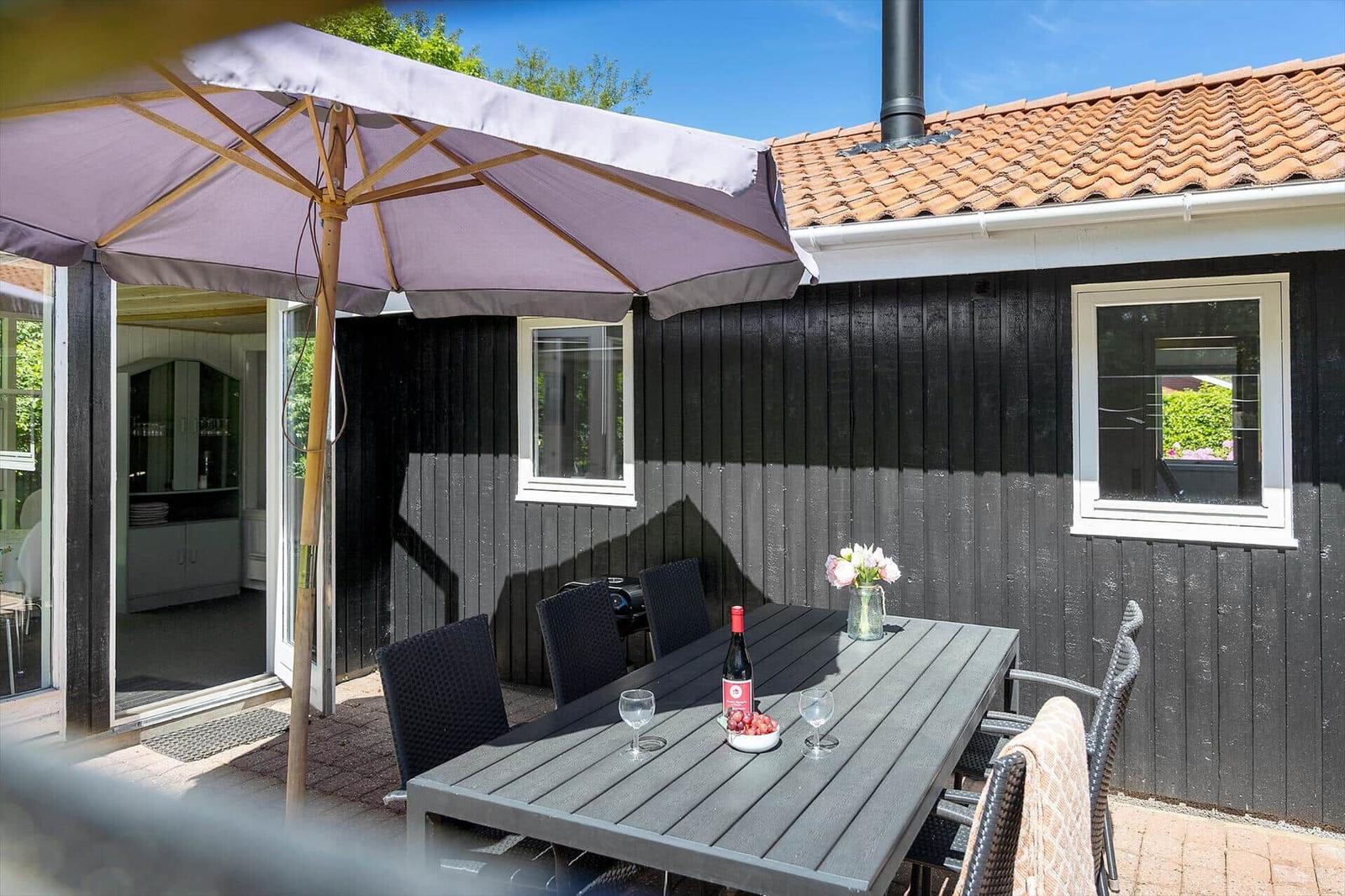 Terrasse mit Tisch, Stühlen und Sonnenschutz vor einem Haus mit schwarzer Fassade.