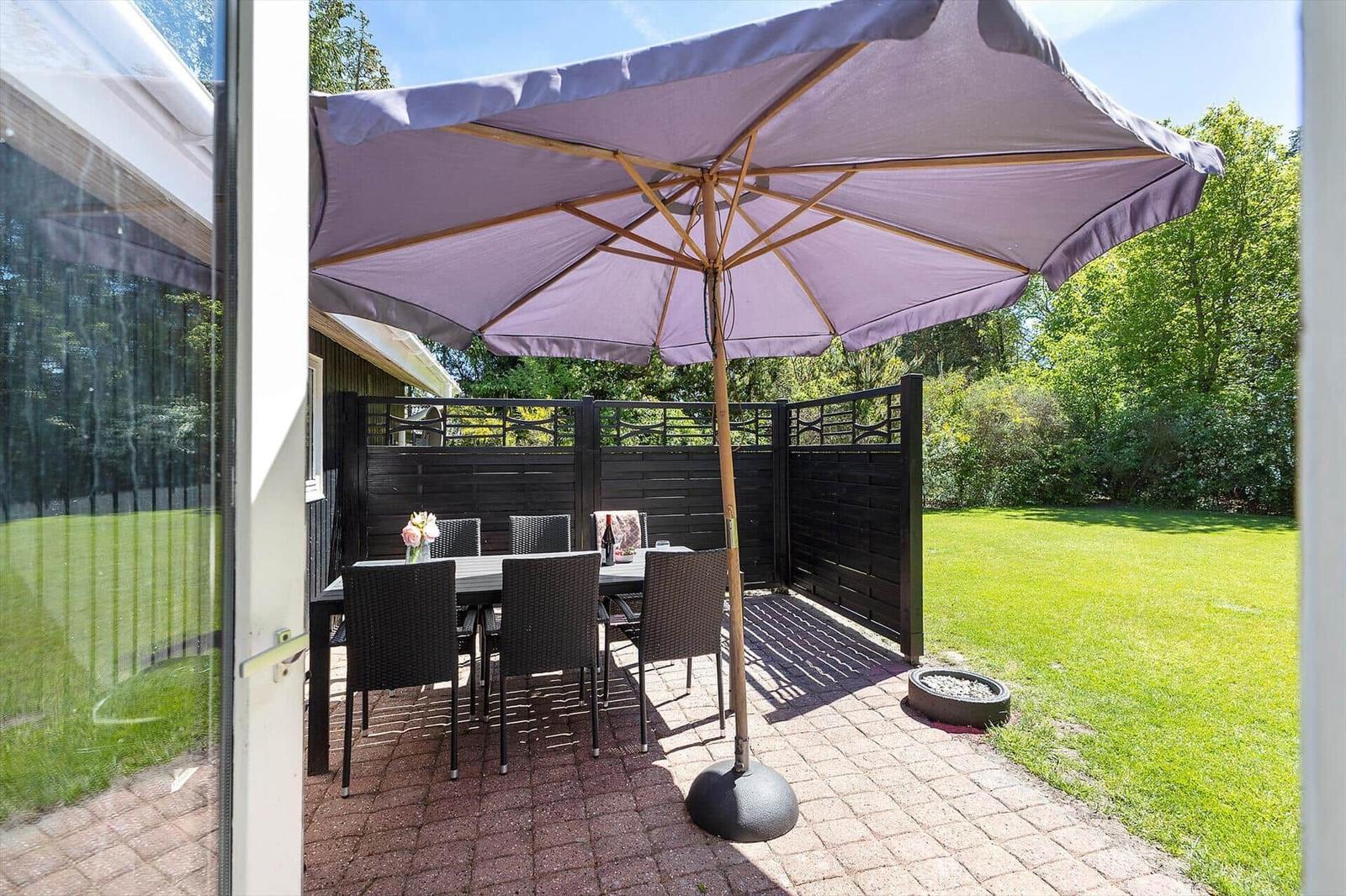 Terrasse mit Tisch, Stühlen und Sonnenschirm. Grünflächen und Zaun im Hintergrund.