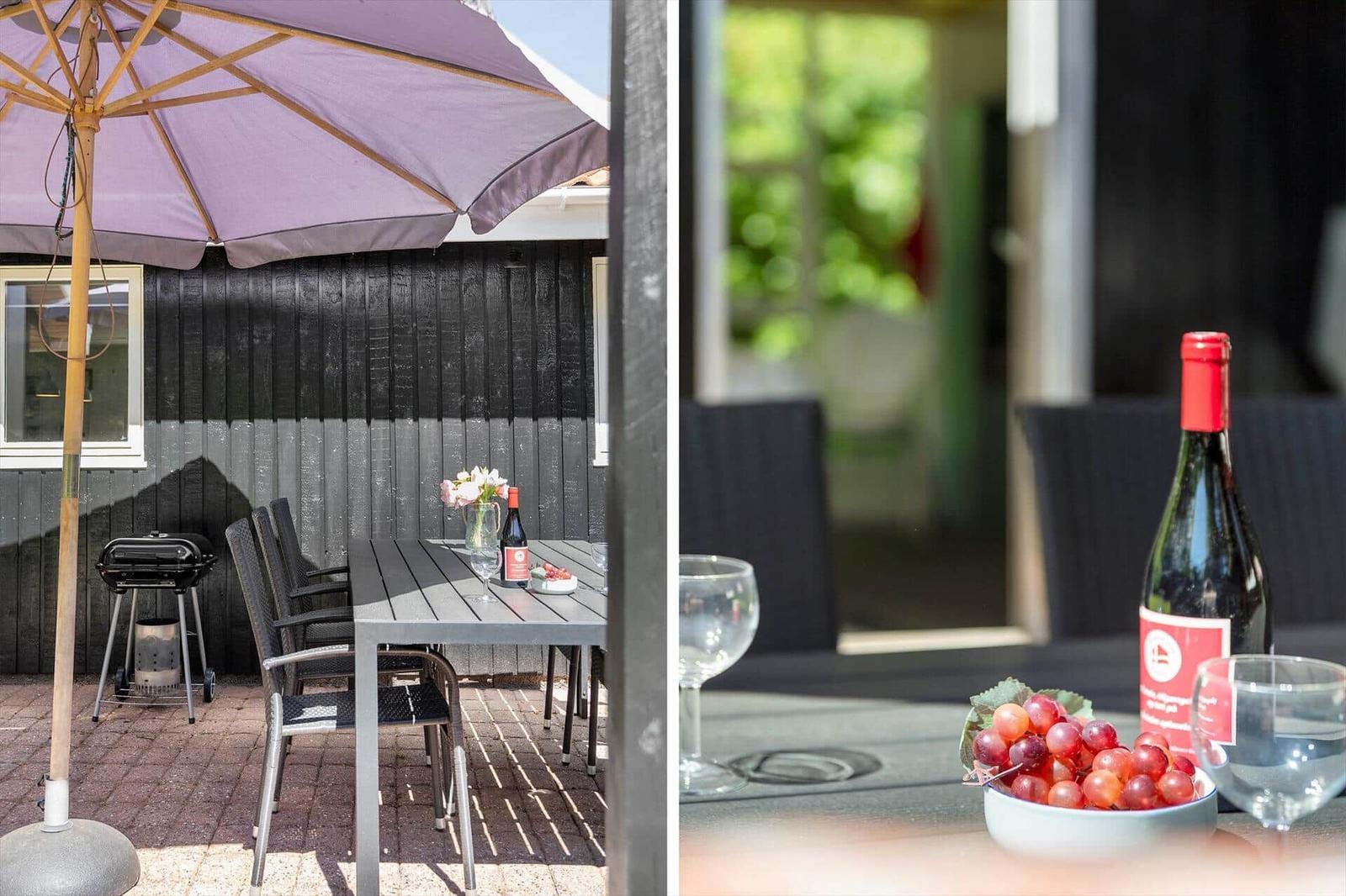 Terrasse mit Tisch, Stühlen, Sonnenschirm und Grill. Auf dem Tisch steht eine Flasche Wein und eine Schale Trauben.