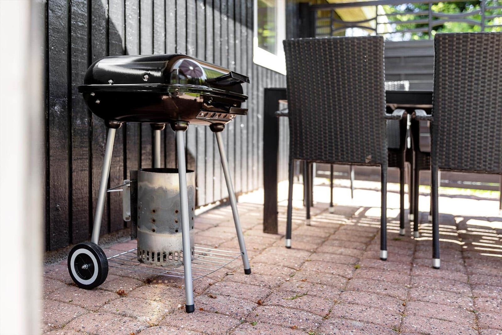 Schwarzer Grill mit Rädern steht auf Terrasse neben Tisch und Stühlen.