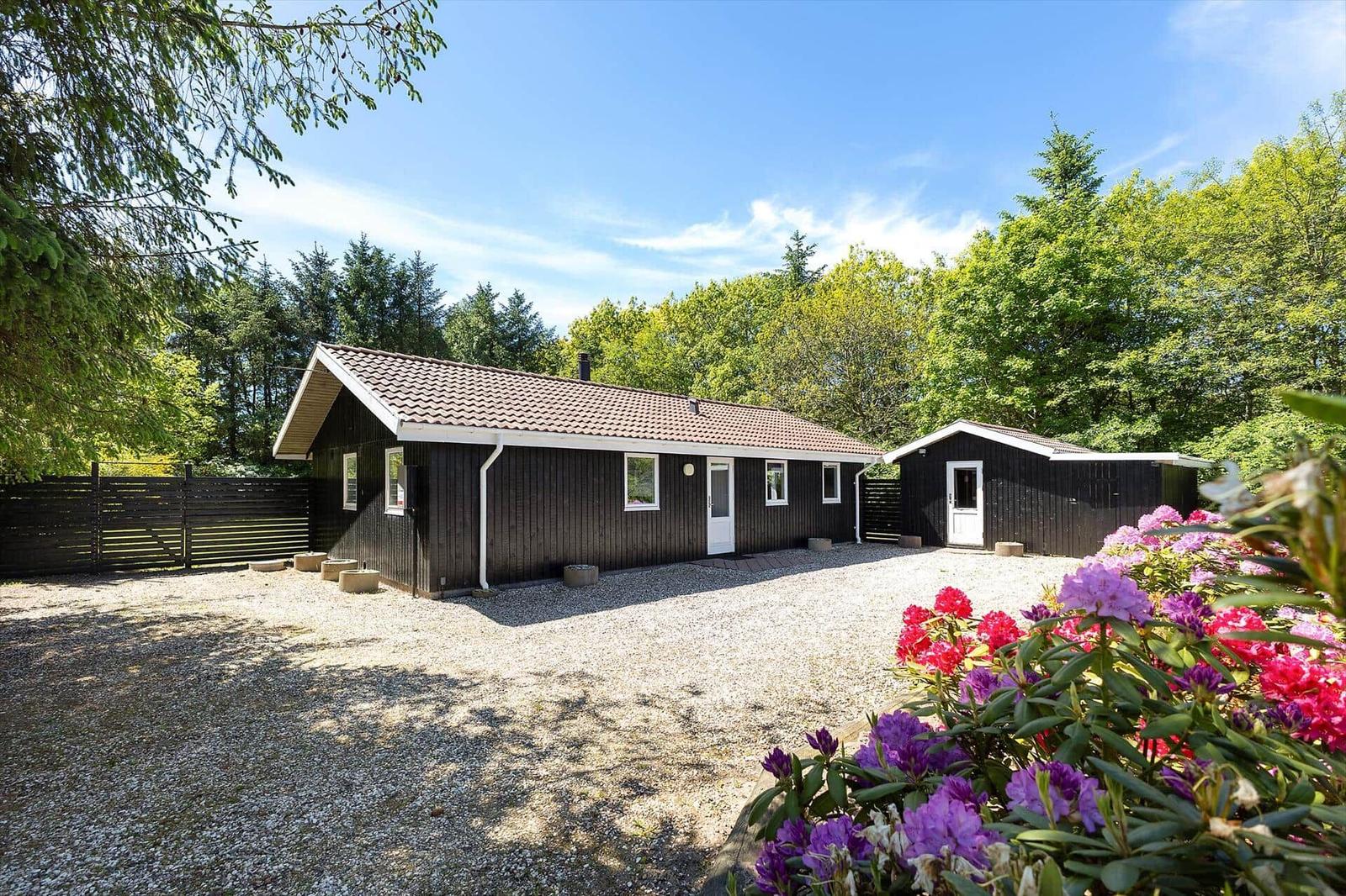 Schwarze Holzhütte mit Garten und Blumen. Umgeben von Bäumen unter blauem Himmel.
