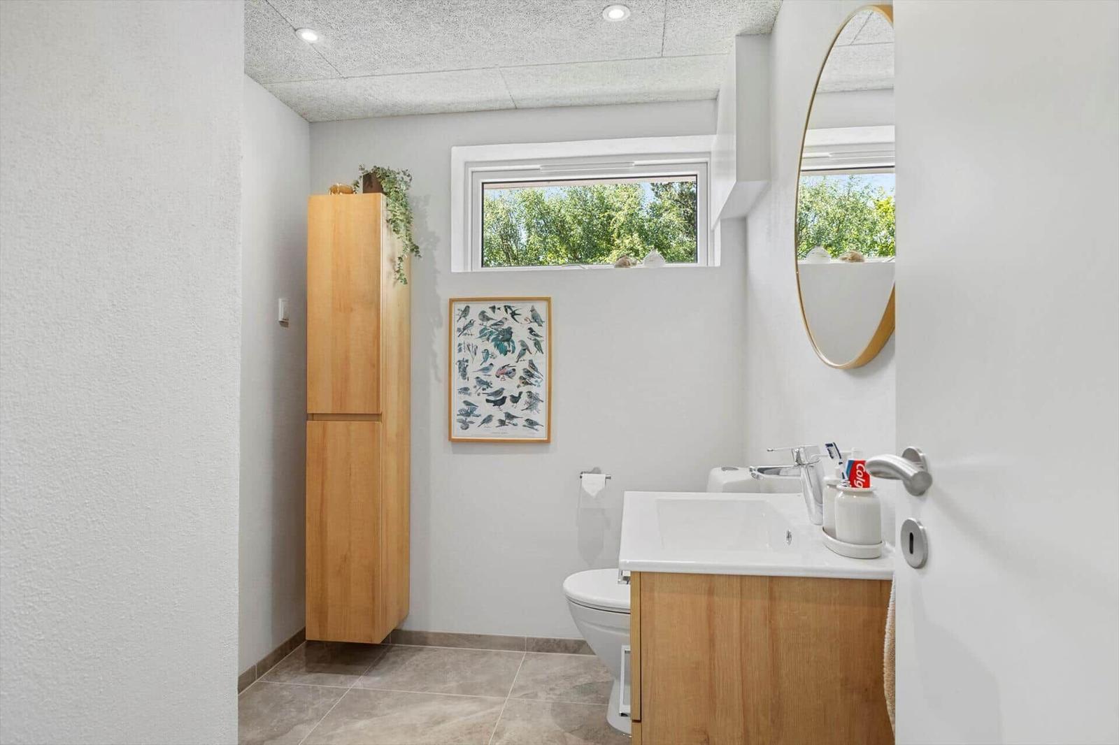 Badezimmer mit Holzschrank, Waschbecken und Fenster mit Blick auf Bäume.