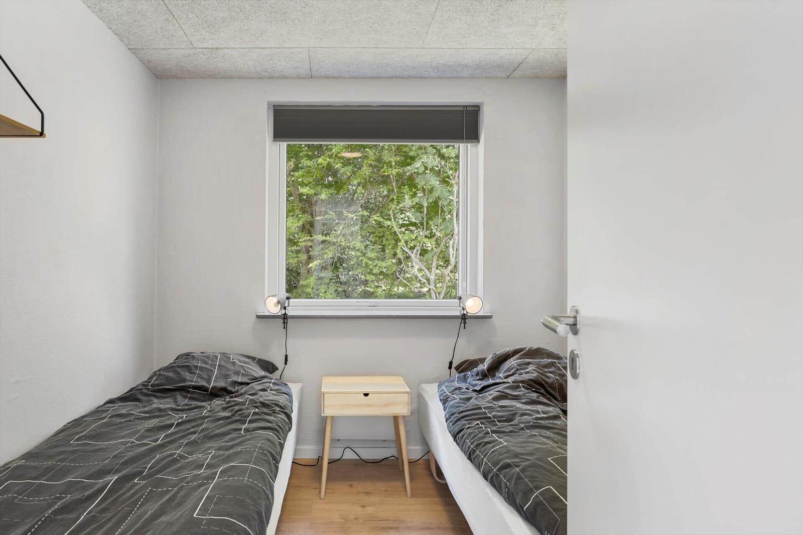 Zwei Einzelbetten mit grauer Bettwäsche, kleiner Nachttisch und Fenster mit Blick auf Bäume.