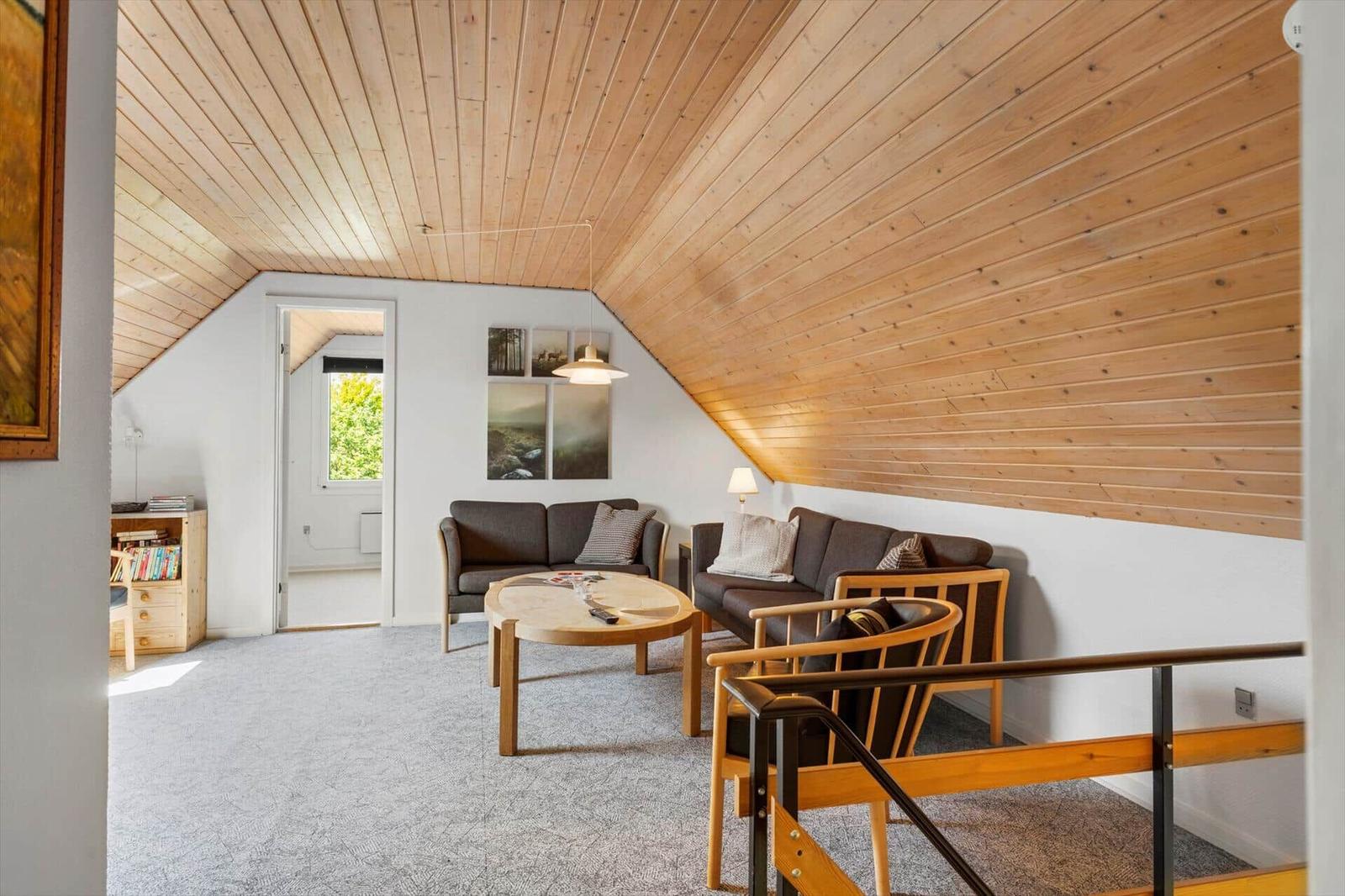 Wohnzimmer mit Holzdecke, Sofa, Tisch und Stühle. Tür führt zu einem weiteren Raum.