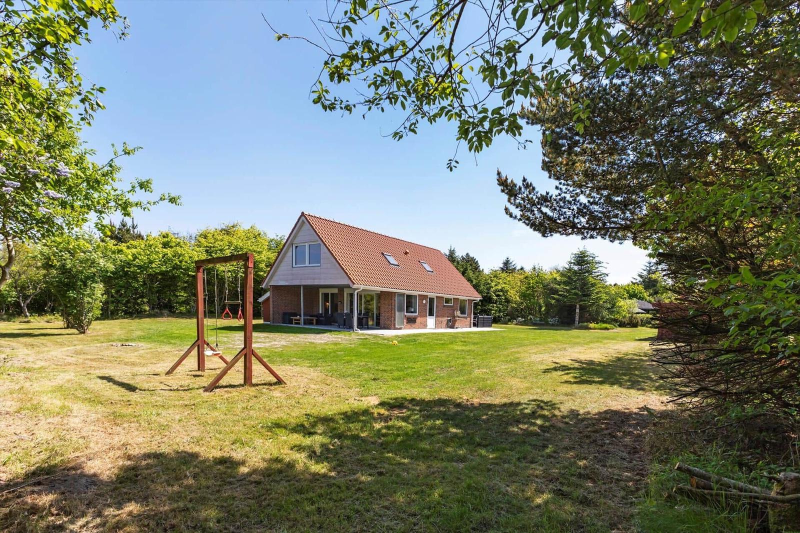 Ein Haus mit rotem Dach und großer Terrasse auf einer grünen Wiese mit Spielplatz.