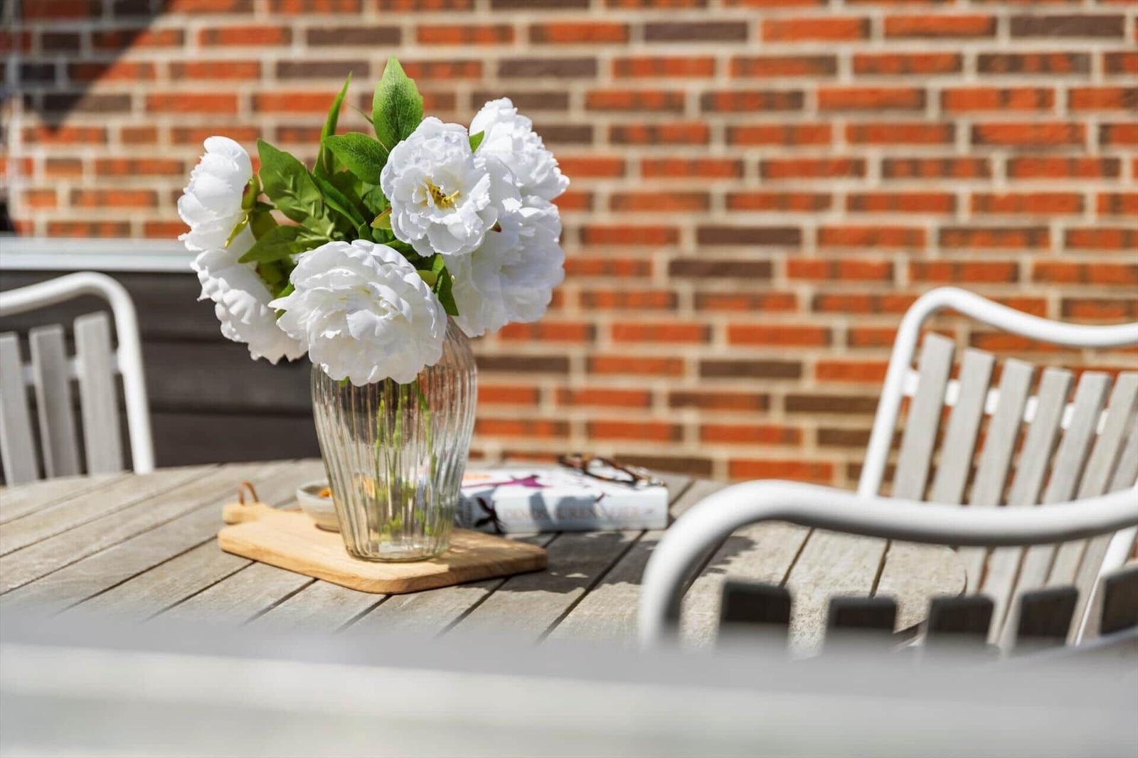 Auf der Terrasse steht ein Tisch mit weißen Blumen in einem Glas. Hintergrund ist eine Backsteinmauer.