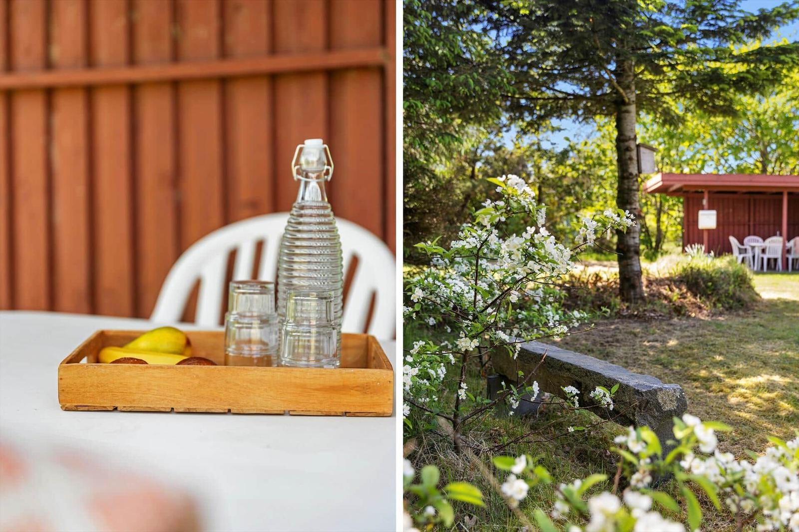 Tisch mit Holzkiste, Gläsern und Obst vor Holzwand. Garten mit Blüten, Steinbank und Pavillon.