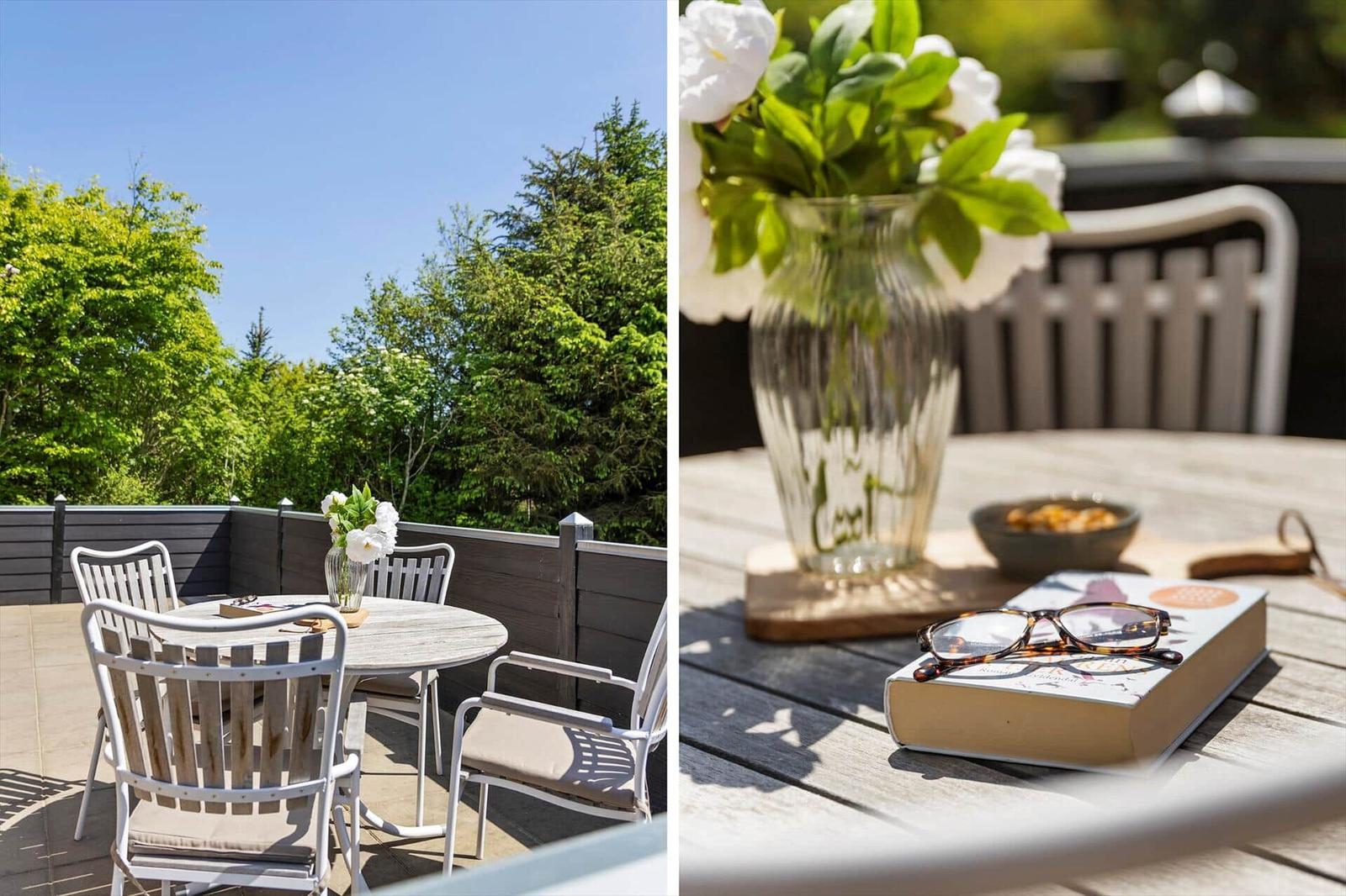 Terrasse mit Tisch und Stühlen, Blumen und Buch. Blick auf grüne Bäume.