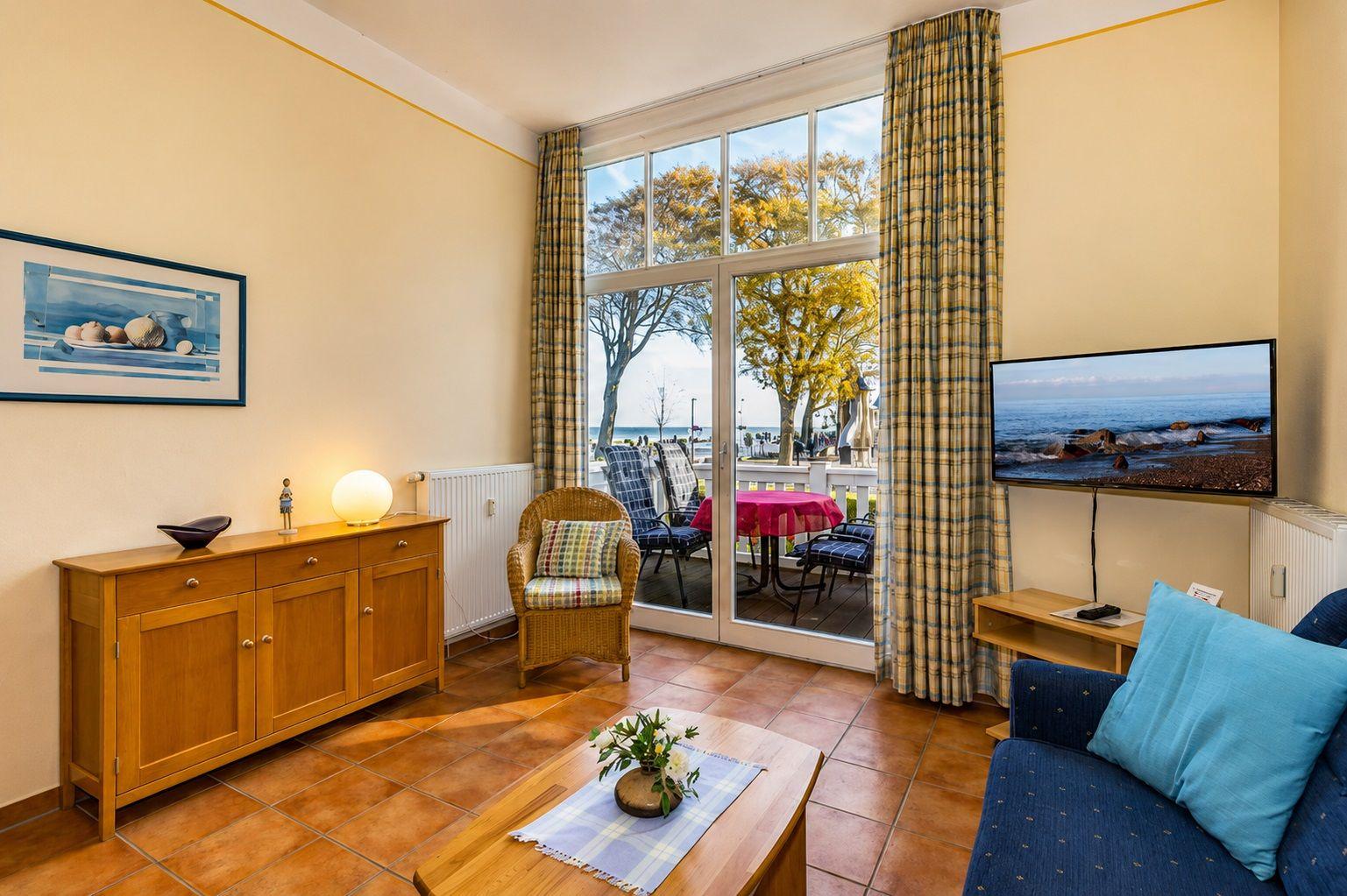 Wohnzimmer mit TV, Sofa und Blick auf den Strand. Durchgangstür führt auf Balkon mit Tisch und Stühlen.
