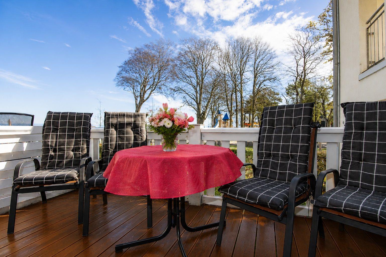 Terrasse mit Tisch, vier Stühlen und Blumenarrangement. Hintergrund: Bäume und Spielplatz.