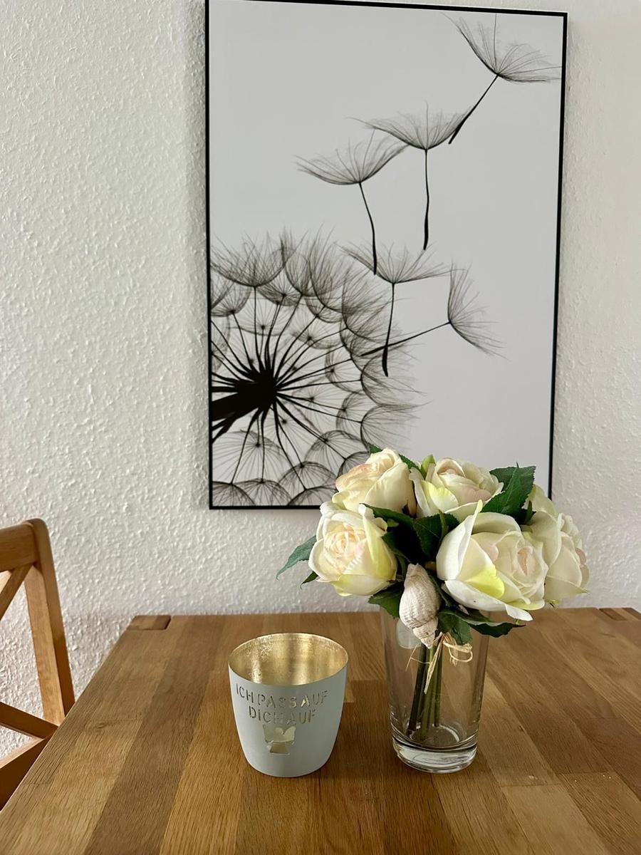 Holztisch mit Blumen in Glas und Kerze vor Wandbild mit Löwenzahn.