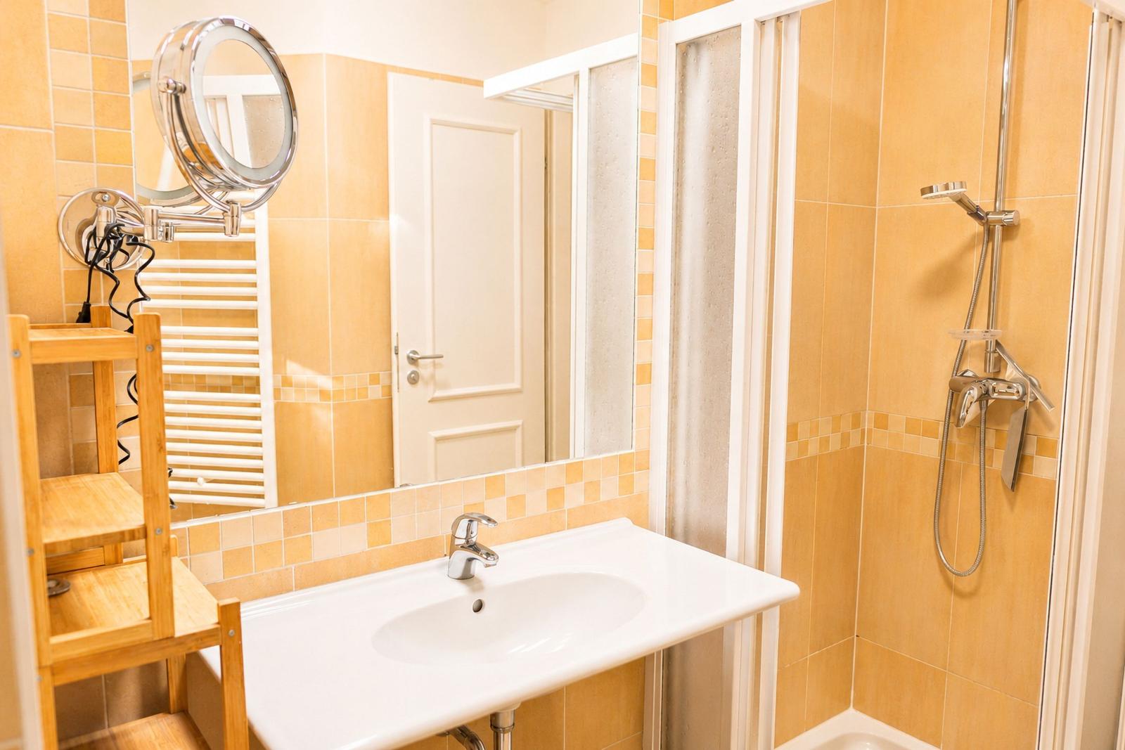 Badezimmer mit Dusche, Waschbecken und Spiegel. Holzregal und Heizung sichtbar.