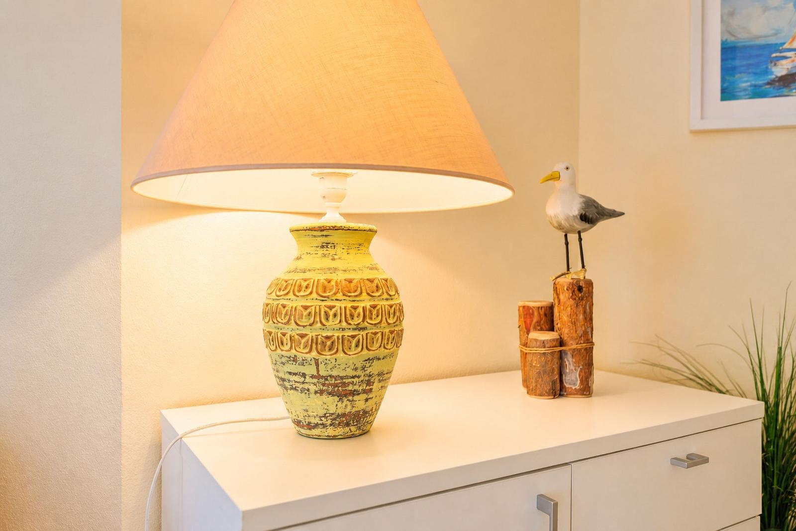 Tischlampe mit gelbem Schirm auf weißen Schrank. Holzdekoration mit Vogelfigur daneben.