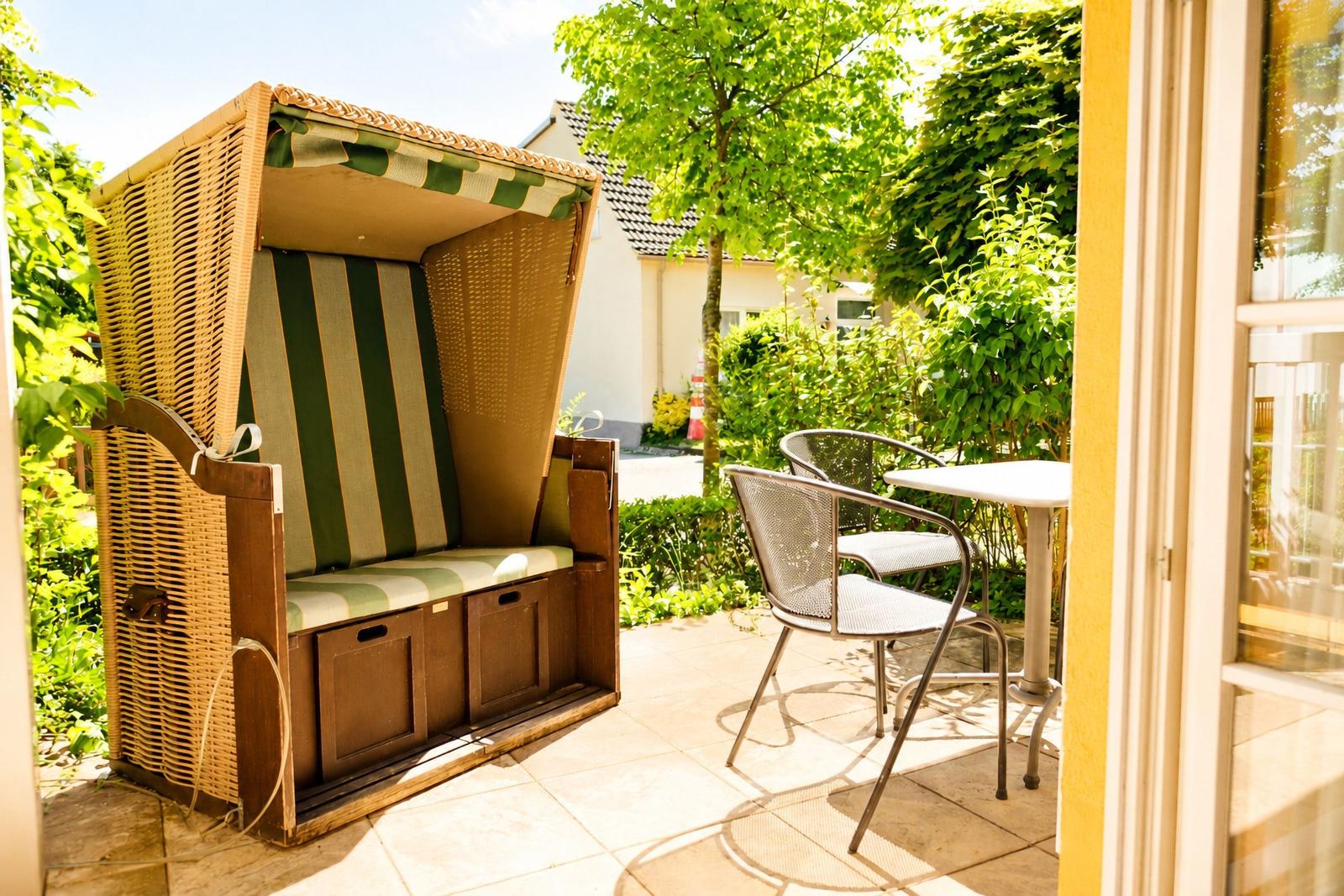 Terrasse mit Strandkorb, Tisch und zwei Stühlen. Grüner Garten im Hintergrund.