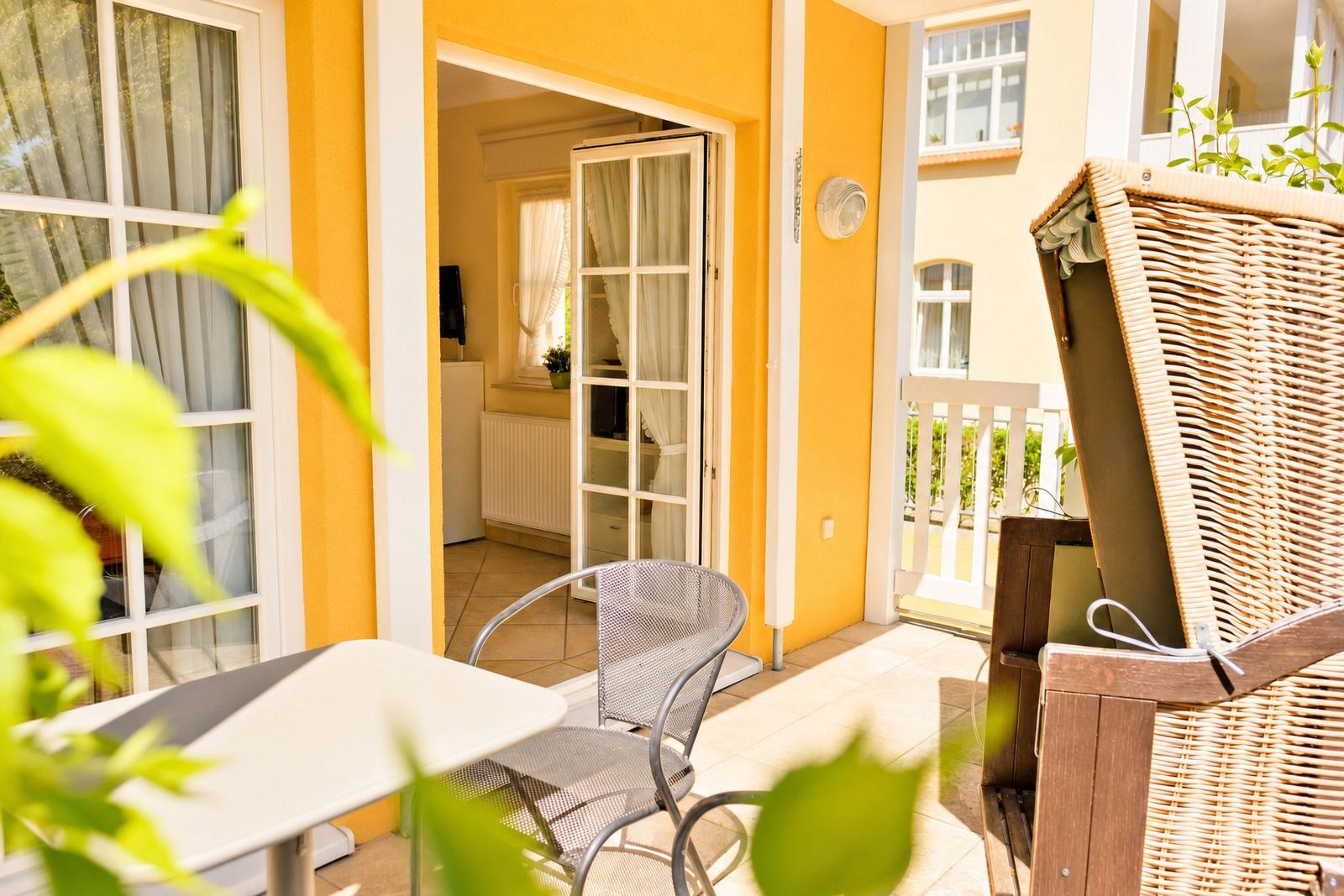 Terrasse mit Tisch, Stühlen und Blick ins Haus. Gelbe Wand, Fenster und Pflanzen.