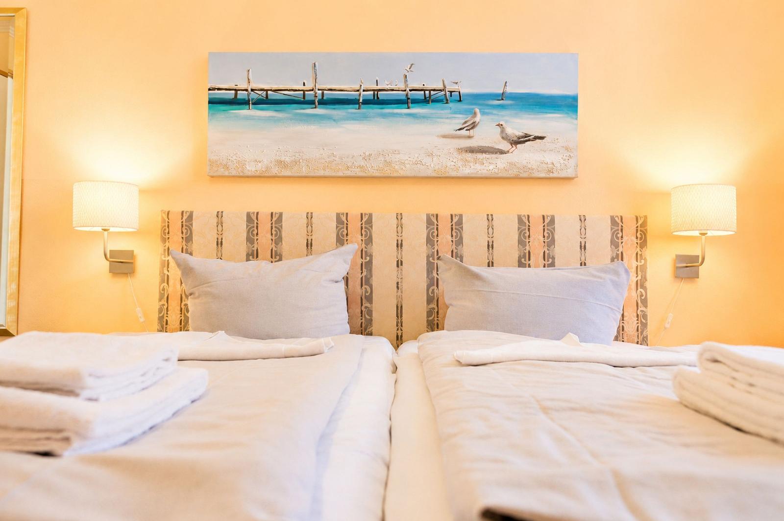 Zwei Betten mit weißen Bettwäsche und Handtüchern. Wandbild zeigt Strand und Pier.