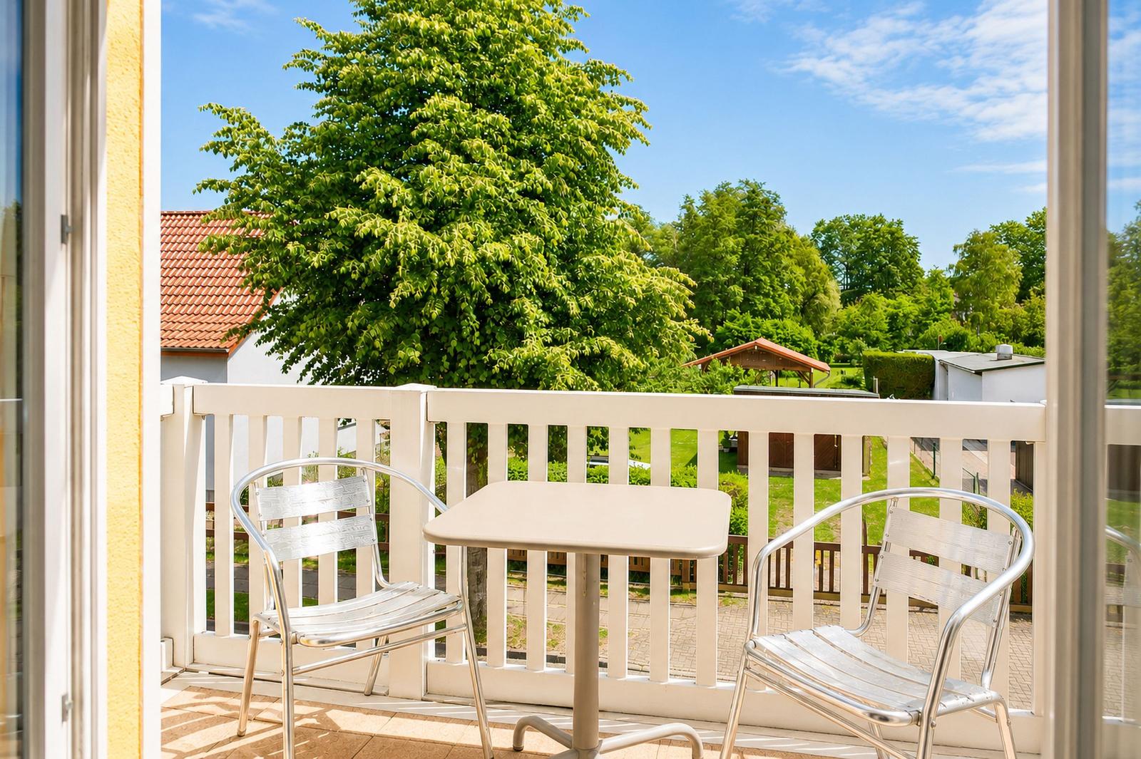 Balkon mit Tisch und zwei Stühlen. Blick auf grüne Bäume und einen Garten.