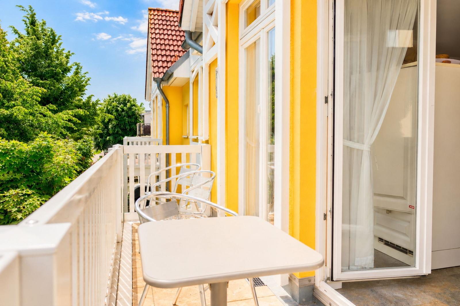 Terrasse mit Tisch und Stühlen, gelbe Fassade und Blick auf grüne Bäume.