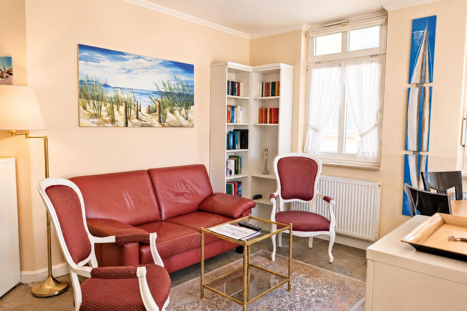Ein Wohnzimmer mit rotem Sofa, weißen Stühlen und Bücherregal. An der Wand hängt ein Strandbild.