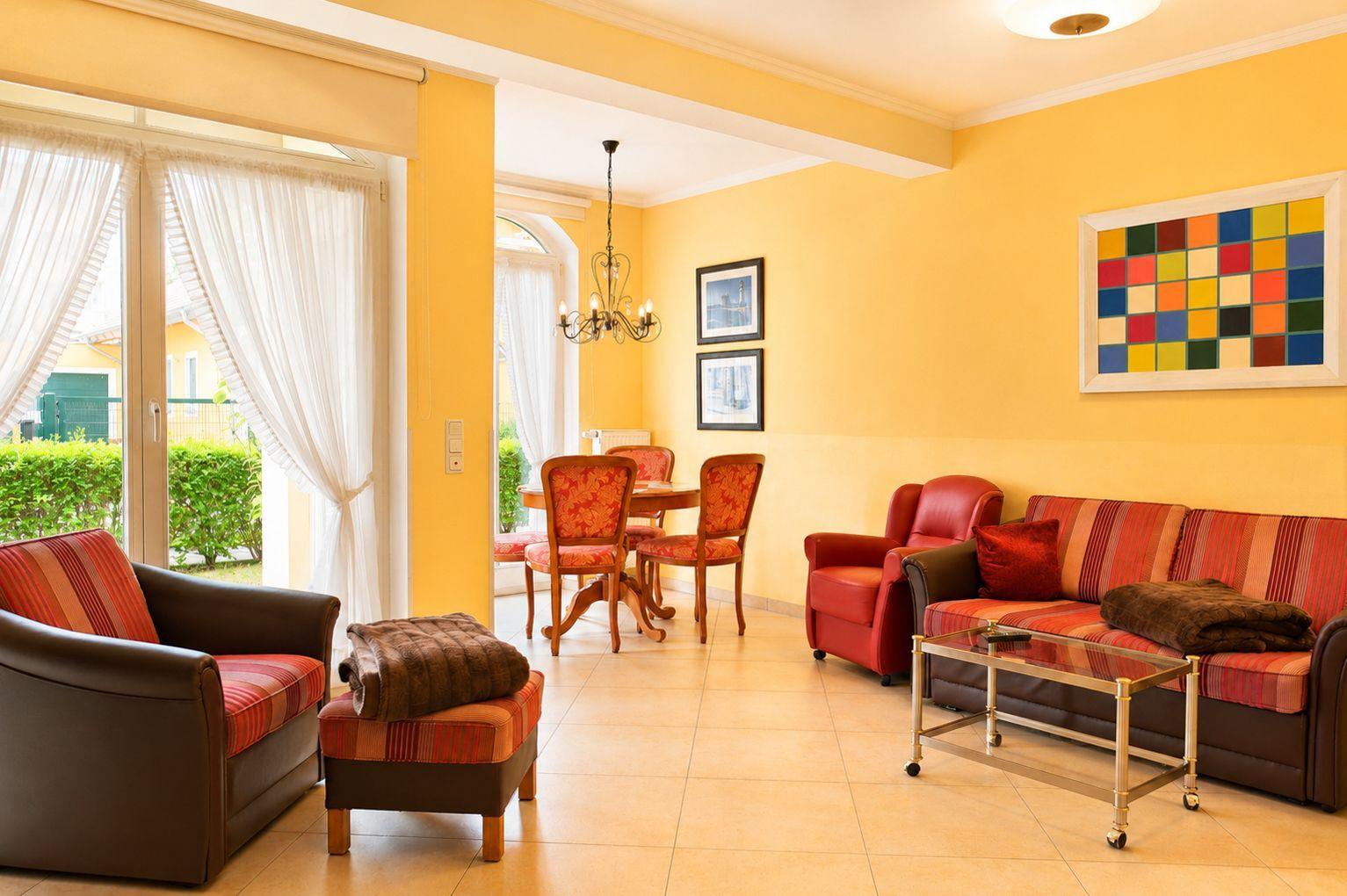 Wohn- und Essbereich mit rotem Sofa, Stühlen und Glas-Tisch. Gelbe Wände und Fenster mit weißen Vorhängen.