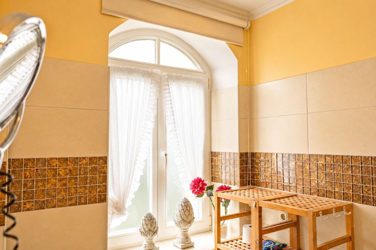 Badezimmer mit gelben Wänden, Holzregal und Fenster mit weißen Vorhängen.