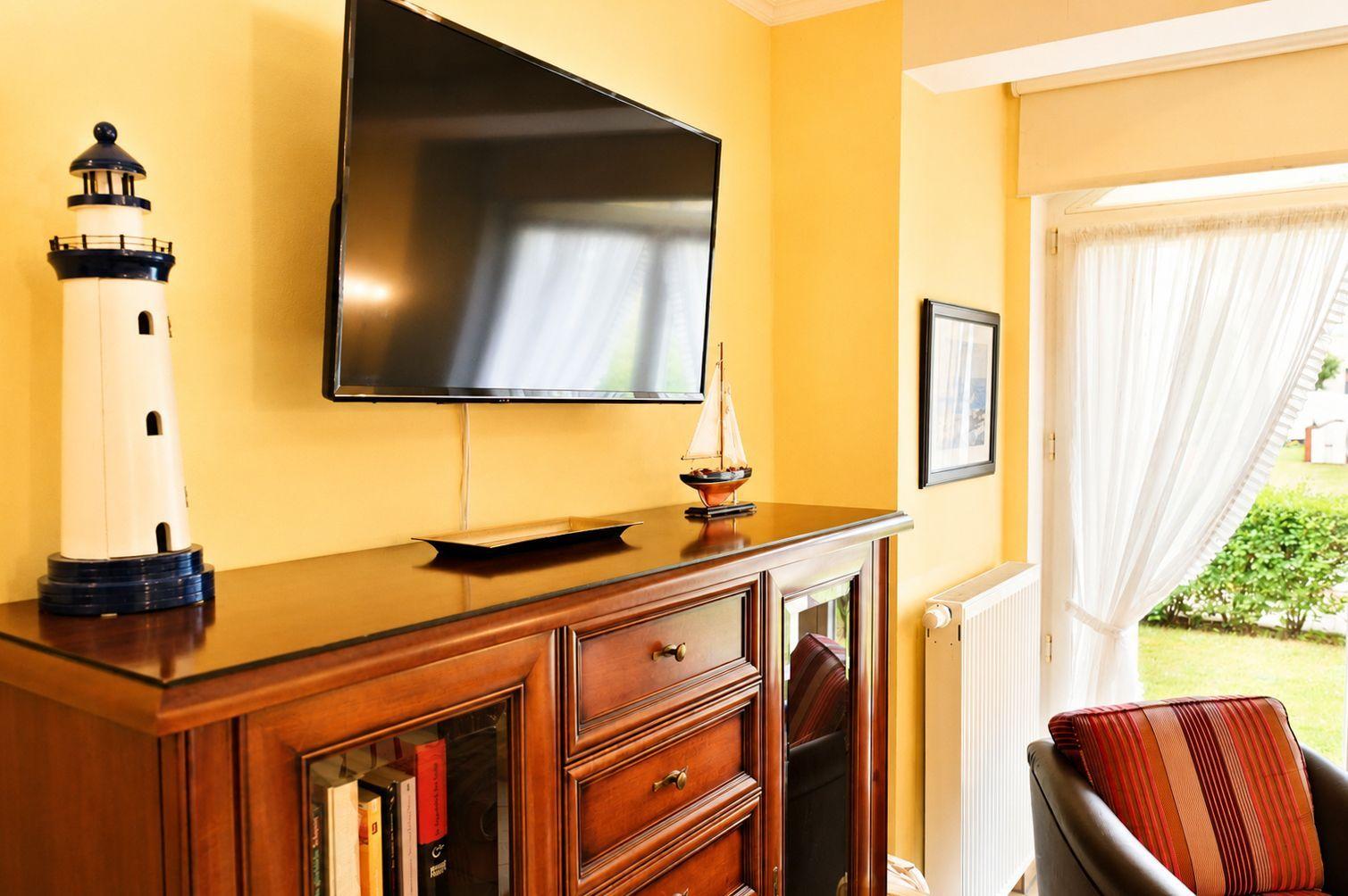 Wohnzimmer mit Fernseher, Holzschrank, Leuchtturm- und Segelschiffmodell sowie Sitzgelegenheit.