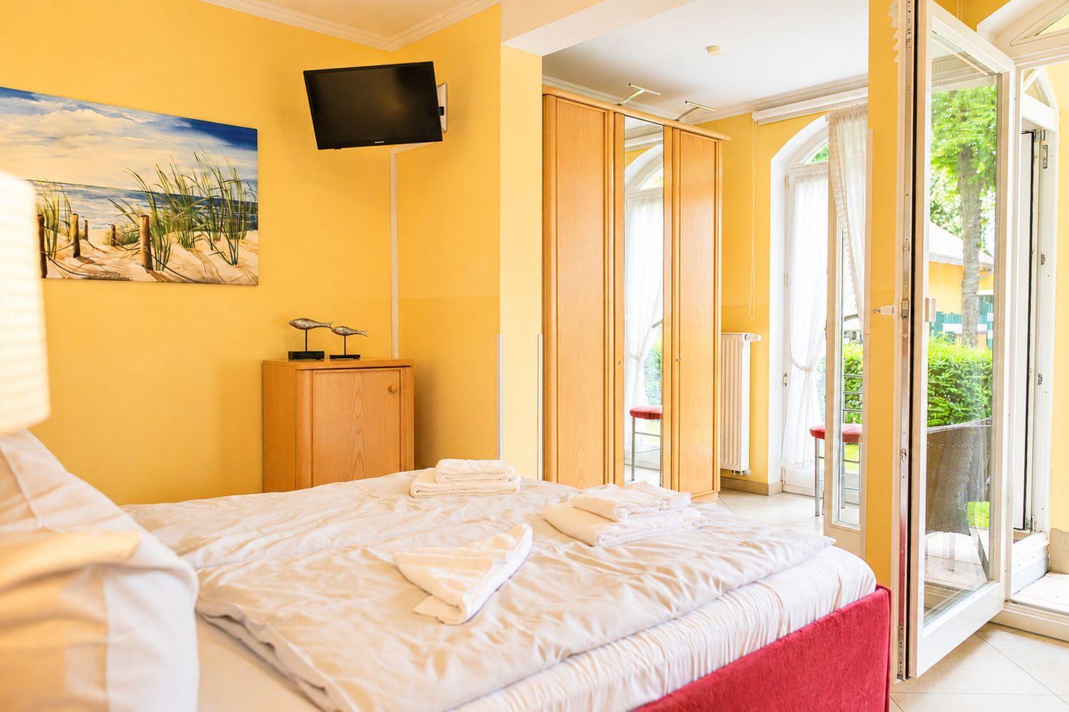 Schlafzimmer mit Bett, TV und Balkonzugang. Gelbe Wände und Holzmöbel.